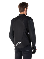 Alpinestars Giacca Impermeabile T-Jaws V4, nero e bianco, vista posteriore, giacca da moto sportiva in tessuto con logo bianco sulla parte bassa della schiena, indossata con guanti e pantaloni neri, con silhouette dalla vestibilità sportiva rivista