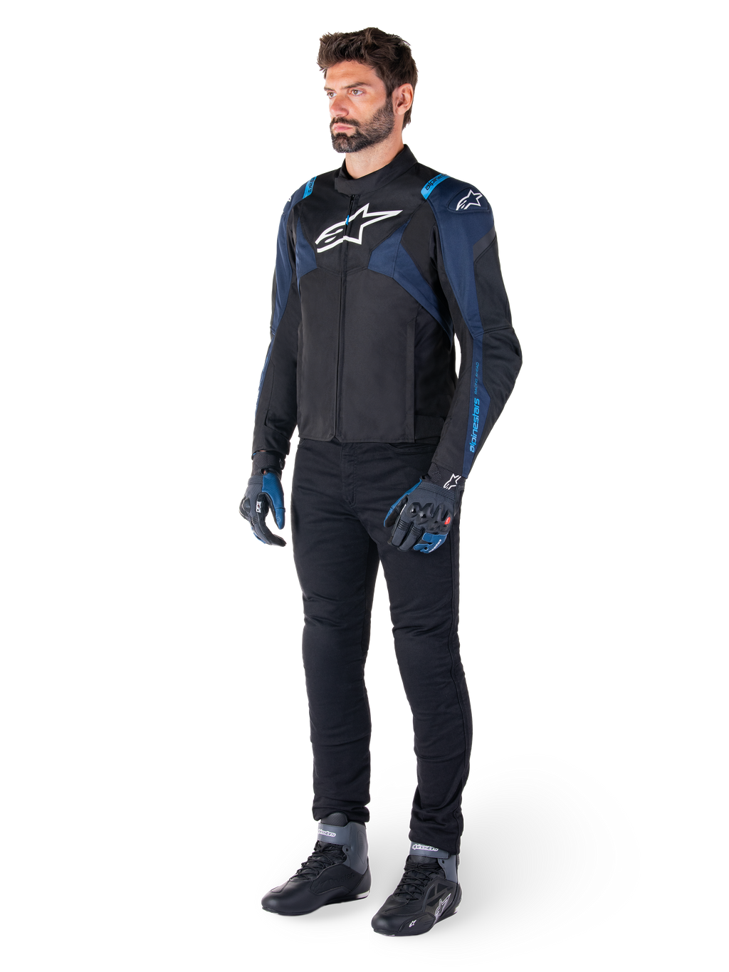 Giacca impermeabile Alpinestars T-Jaws V4 in nero, blu scuro e blu Blithe, indossata da un modello con pantaloni da moto neri e stivali da guida, con logo Alpinestars bianco sul petto e accenti blu sulle spalle, abbigliamento tecnico sport-fit