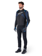 Giacca impermeabile Alpinestars T-Jaws V4 in nero, blu scuro e blu Blithe, indossata da un modello con pantaloni da moto neri e stivali da guida, con logo Alpinestars bianco sul petto e accenti blu sulle spalle, abbigliamento tecnico sport-fit