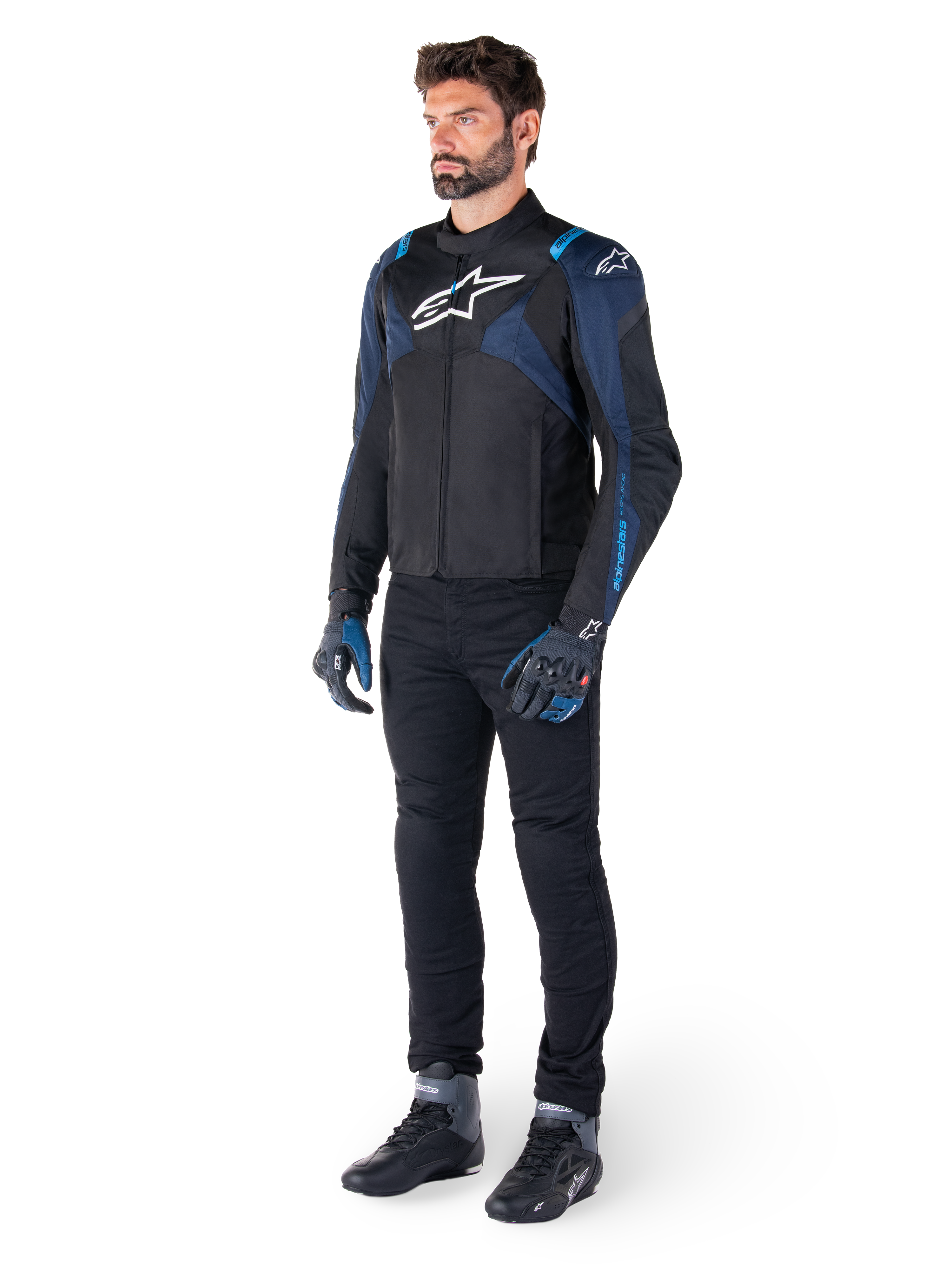 Giacca impermeabile Alpinestars T-Jaws V4 in nero, blu scuro e blu Blithe, indossata da un modello con pantaloni da moto neri e stivali da guida, con logo Alpinestars bianco sul petto e accenti blu sulle spalle, abbigliamento tecnico sport-fit