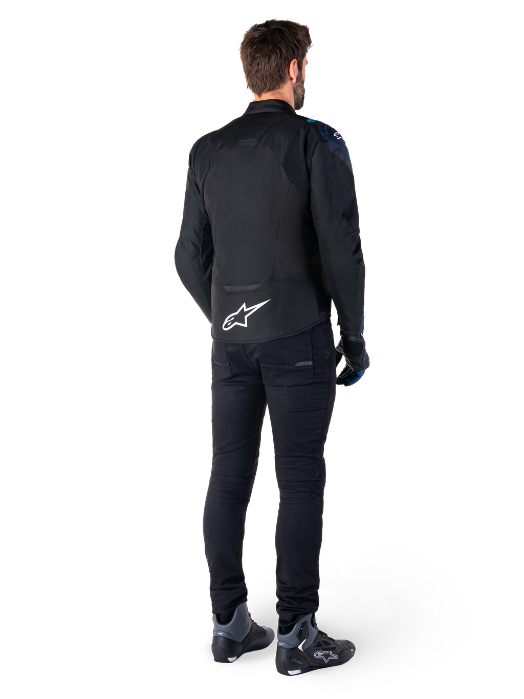 Vista posteriore di un uomo che indossa la giacca Alpinestars T-Jaws V4 Waterproof in nero, blu scuro e blu Blithe, caratterizzata da un evidente logo Astar bianco sulla parte bassa della schiena, abbinata a pantaloni neri e scarpe da moto.