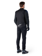 Vista posteriore di un uomo che indossa la giacca Alpinestars T-Jaws V4 Waterproof in nero, blu scuro e blu Blithe, caratterizzata da un evidente logo Astar bianco sulla parte bassa della schiena, abbinata a pantaloni neri e scarpe da moto.