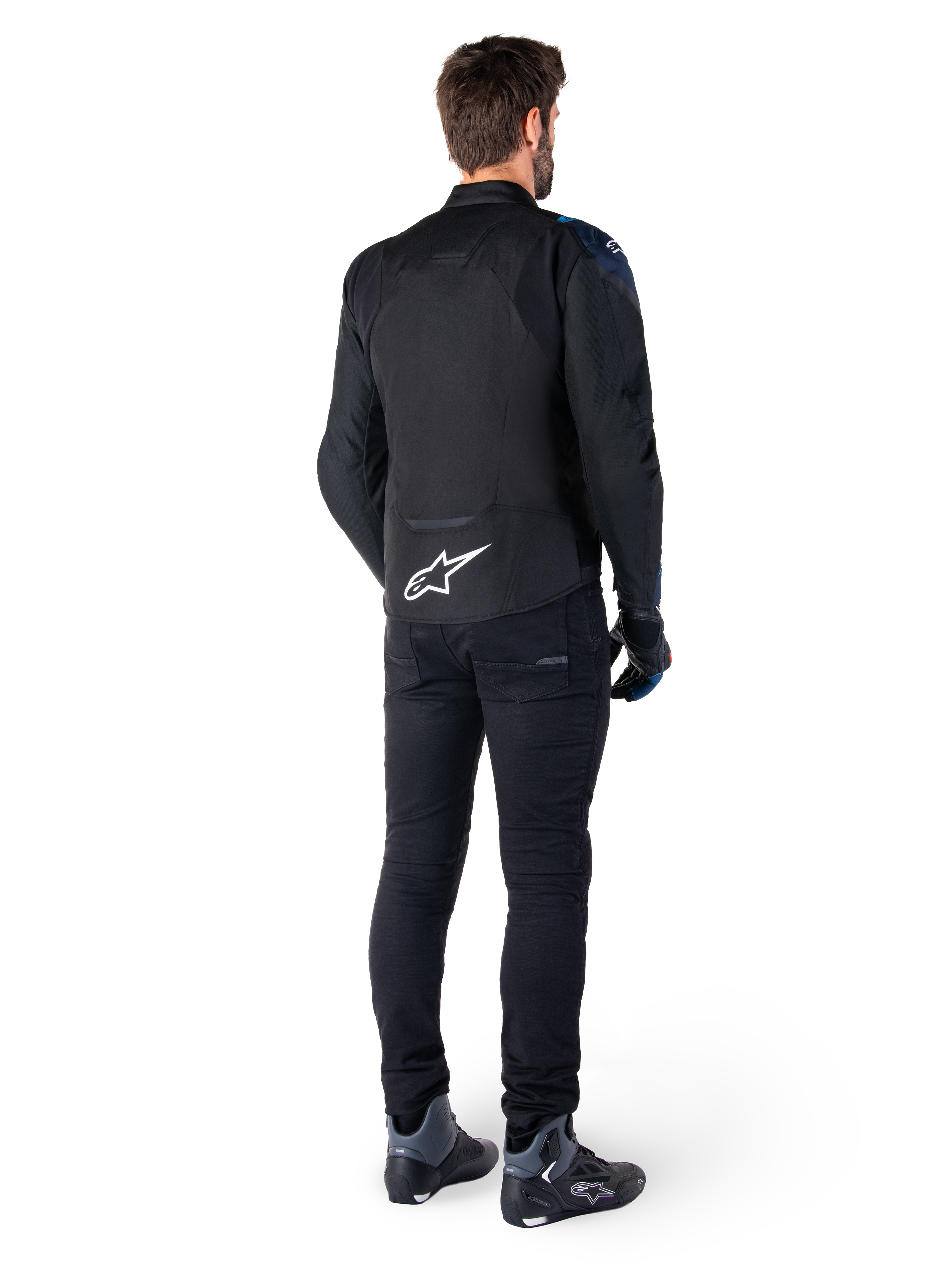 Vista posteriore di un uomo che indossa la giacca Alpinestars T-Jaws V4 Waterproof in nero, blu scuro e blu Blithe, caratterizzata da un evidente logo Astar bianco sulla parte bassa della schiena, abbinata a pantaloni neri e scarpe da moto.