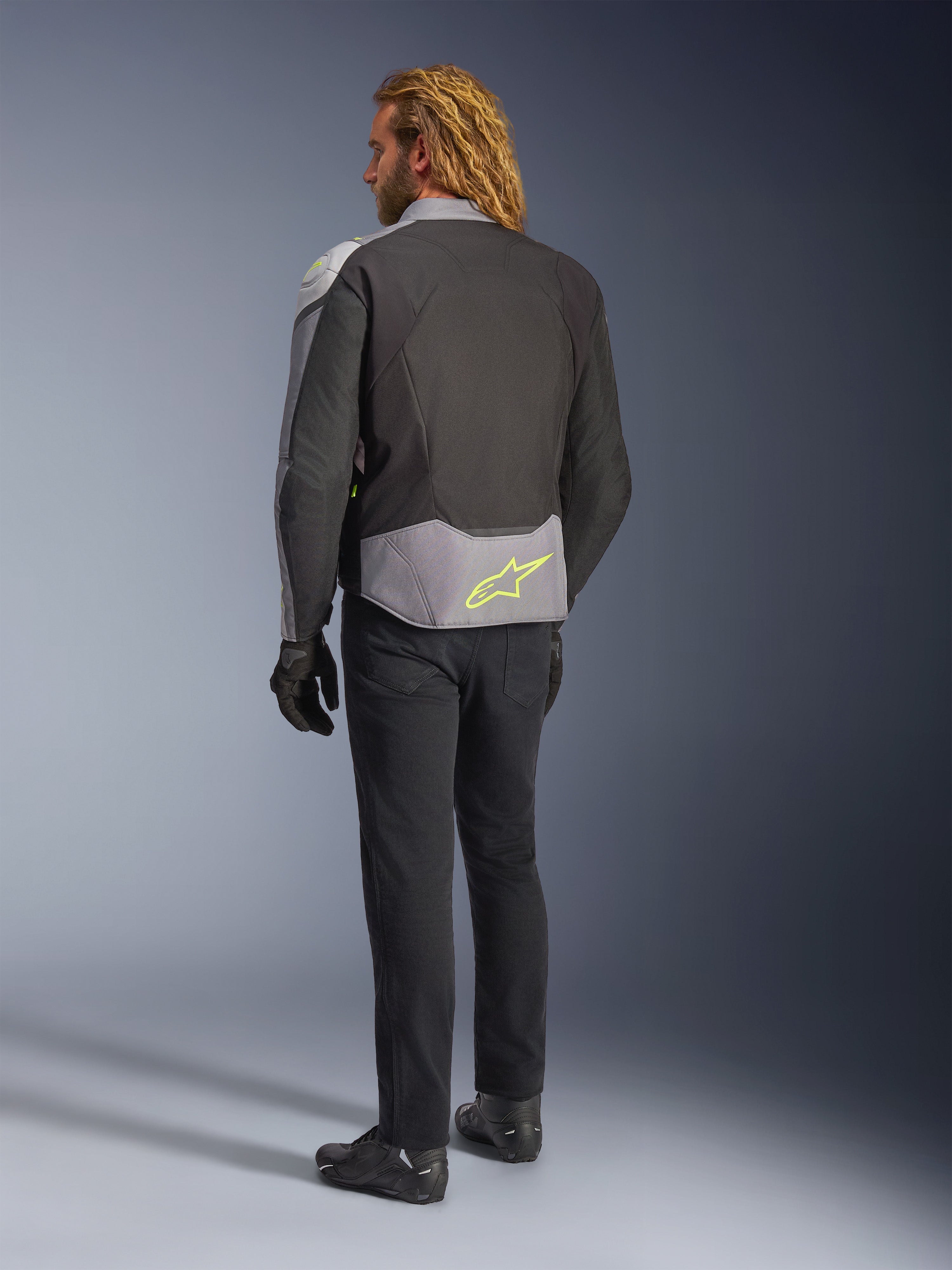 Giacca impermeabile Alpinestars T-Jaws V4, vista posteriore, Black Ice Gray e Dark Gray, con logo Alpinestars giallo neon sulla parte bassa della schiena, indossata con pantaloni neri e guanti da moto, abbigliamento da guida sportivo