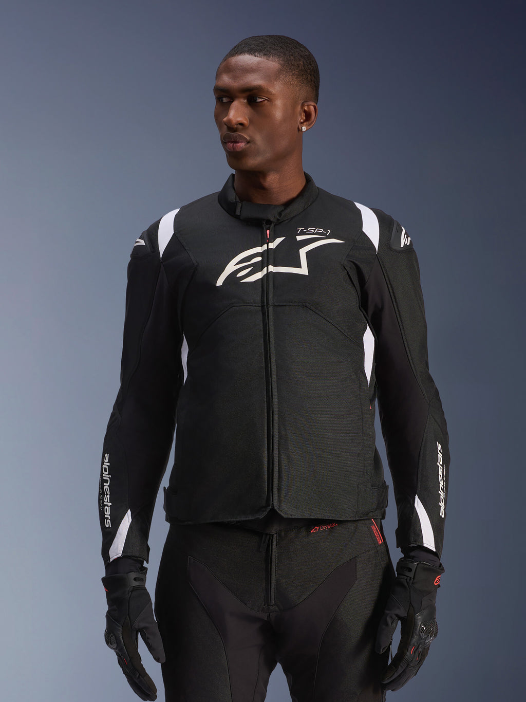 Giacca impermeabile Alpinestars T-SP 1 V2, giacca moto in tessuto sportivo, nera e bianca con sottili dettagli rossi, indossata da un modello uomo con guanti e pantaloni coordinati, con logo Alpinestars bianco sul petto, marchio T-SP-1 e silhouette protettiva per la guida su strada
