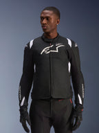 Giacca impermeabile Alpinestars T-SP 1 V2, giacca moto in tessuto sportivo, nera e bianca con sottili dettagli rossi, indossata da un modello uomo con guanti e pantaloni coordinati, con logo Alpinestars bianco sul petto, marchio T-SP-1 e silhouette protettiva per la guida su strada