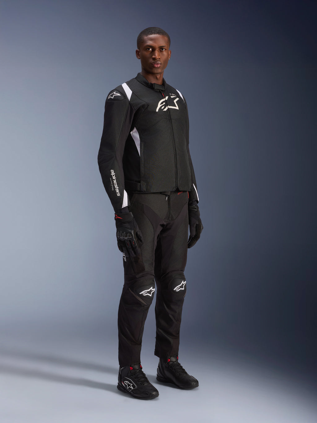 Modello che indossa la giacca impermeabile Alpinestars T-SP 1 V2 in nero e bianco, abbinata a pantaloni da moto, guanti e stivali neri coordinati. Scatto in studio a figura intera di una giacca sportiva in tessuto con loghi bianchi e silhouette sartoriale 3D.