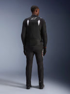 Giacca impermeabile Alpinestars T-SP 1 V2, nero e bianco, vista posteriore di un pilota che indossa la giacca con pantaloni e stivali da moto neri coordinati, con logo A-star bianco sulla parte superiore della schiena e inserti riflettenti sulle spalle, abbigliamento tecnico sportivo in tessuto