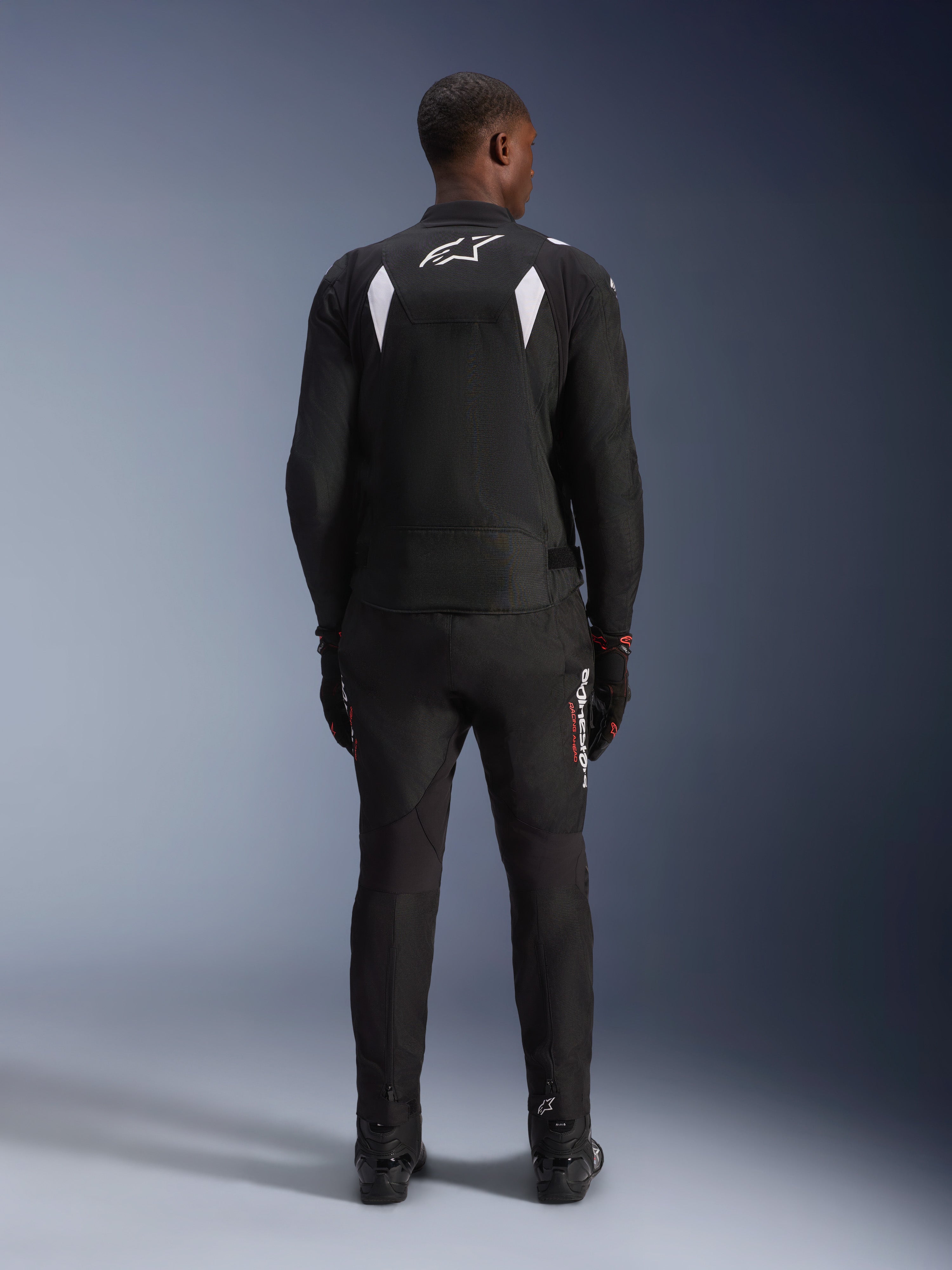 Giacca impermeabile Alpinestars T-SP 1 V2, nero e bianco, vista posteriore di un pilota che indossa la giacca con pantaloni e stivali da moto neri coordinati, con logo A-star bianco sulla parte superiore della schiena e inserti riflettenti sulle spalle, abbigliamento tecnico sportivo in tessuto