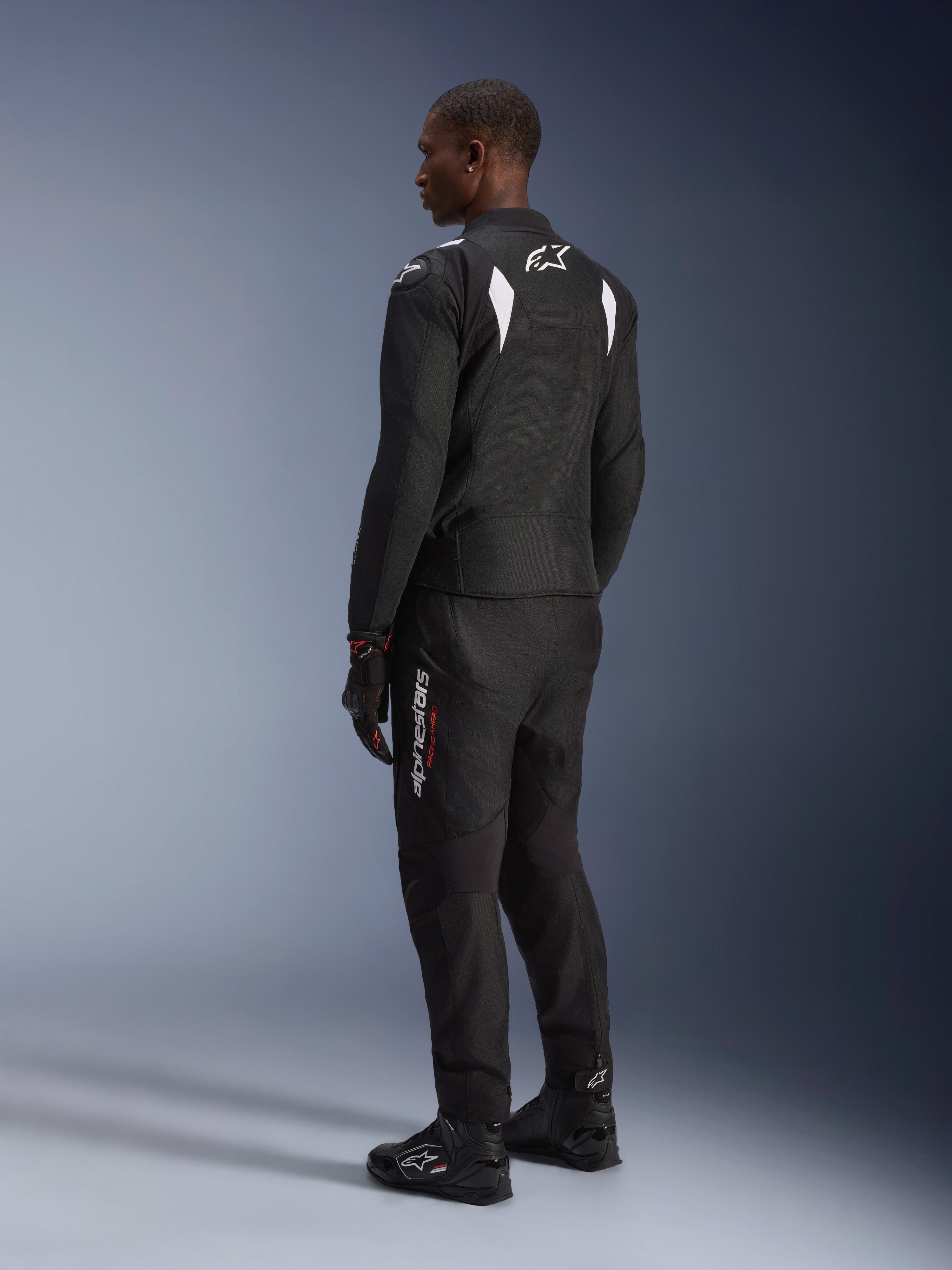 Alpinestars Giacca Impermeabile T-SP 1 V2, nero e bianco, vista posteriore e laterale su modello, indossata con pantaloni da moto, guanti e stivali neri coordinati, completo da moto sportivo in tessuto per guida su strada