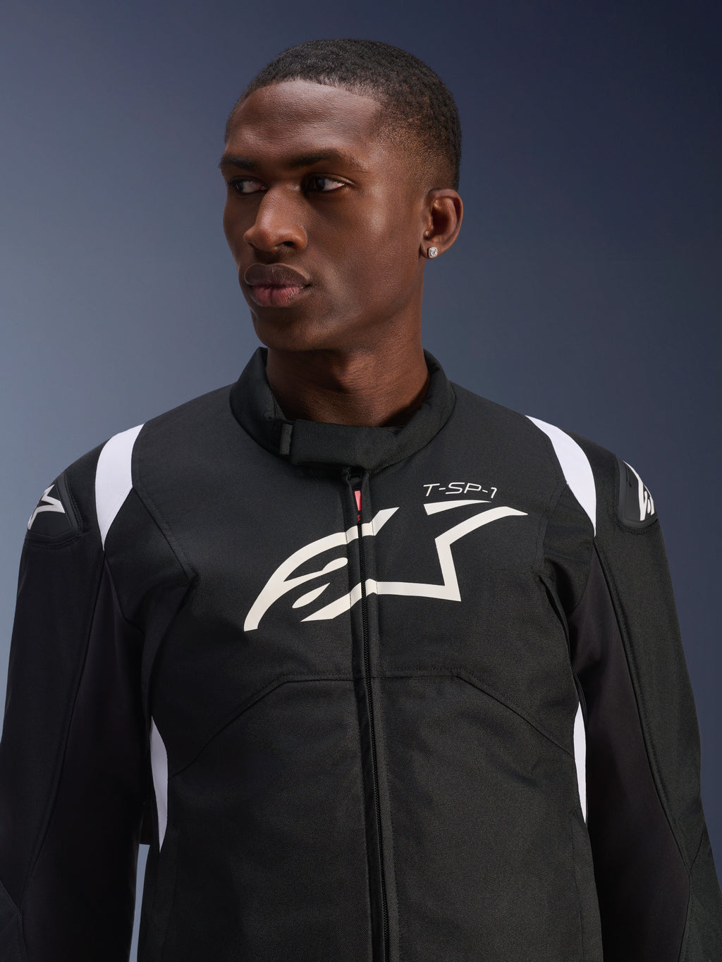 Giacca impermeabile Alpinestars T-SP 1 V2 indossata da un modello, giacca da moto in tessuto bianco e nero, grande logo Alpinestars bianco e marchio T-SP-1 sul petto, design sportivo con slider protettivi sulle spalle
