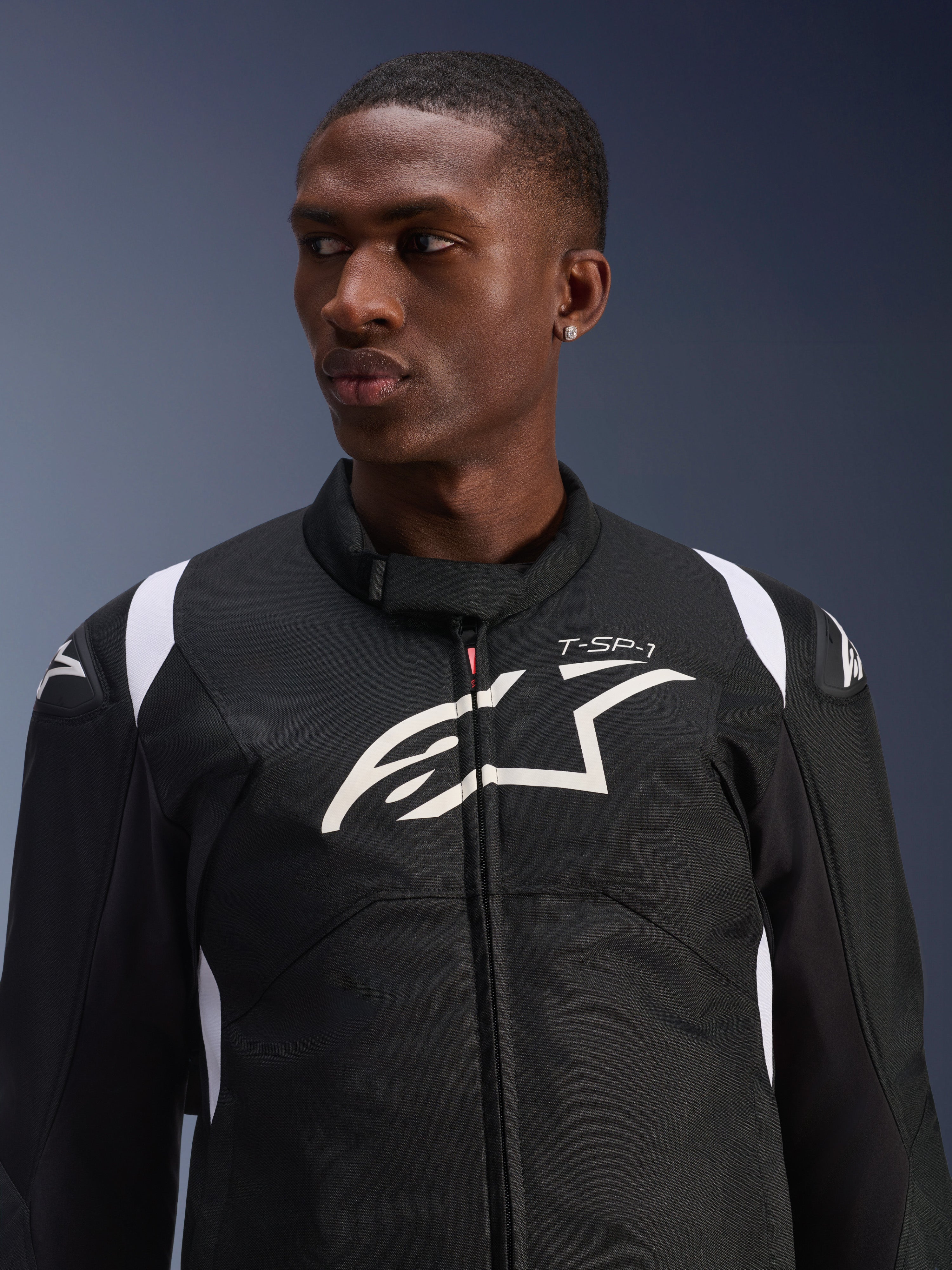 Giacca impermeabile Alpinestars T-SP 1 V2 indossata da un modello, giacca da moto in tessuto bianco e nero, grande logo Alpinestars bianco e marchio T-SP-1 sul petto, design sportivo con slider protettivi sulle spalle