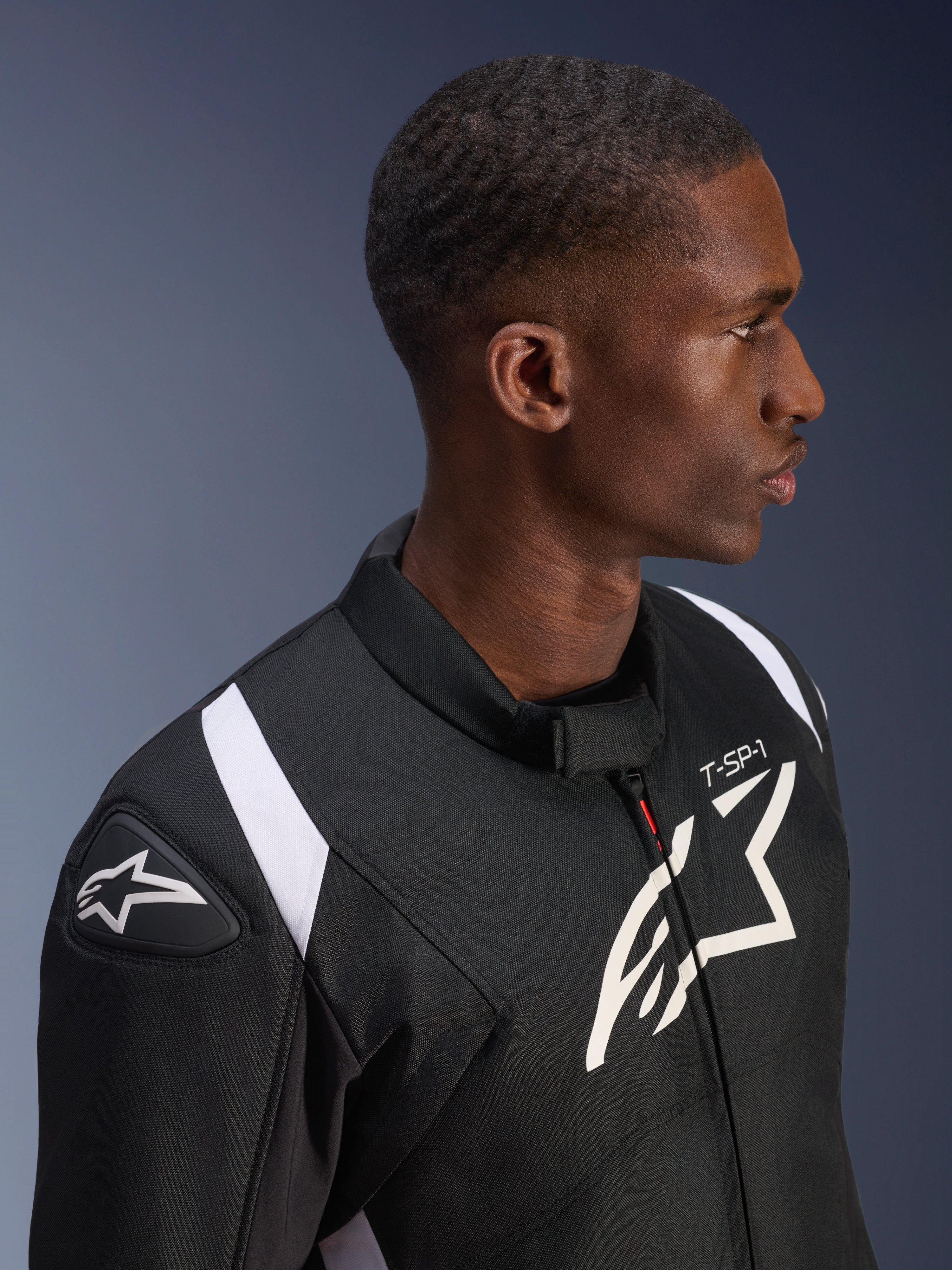 Giacca Impermeabile Alpinestars T-SP 1 V2, nera e bianca, scatto di profilo di un modello che indossa la giacca, con il logo Alpinestars sullo slider della spalla e sul petto, marchio 'T-SP-1' e accenti bianchi sulle spalle.