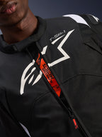 Giacca impermeabile Alpinestars T-SP 1 V2, nero e bianco, con un grande logo bianco sul petto e la scritta 'T-SP-1', patta interna arancione con branding 'Alpinestars Protects' e 'Tech-Air Ready', abbigliamento tecnico da moto in tessuto resistente