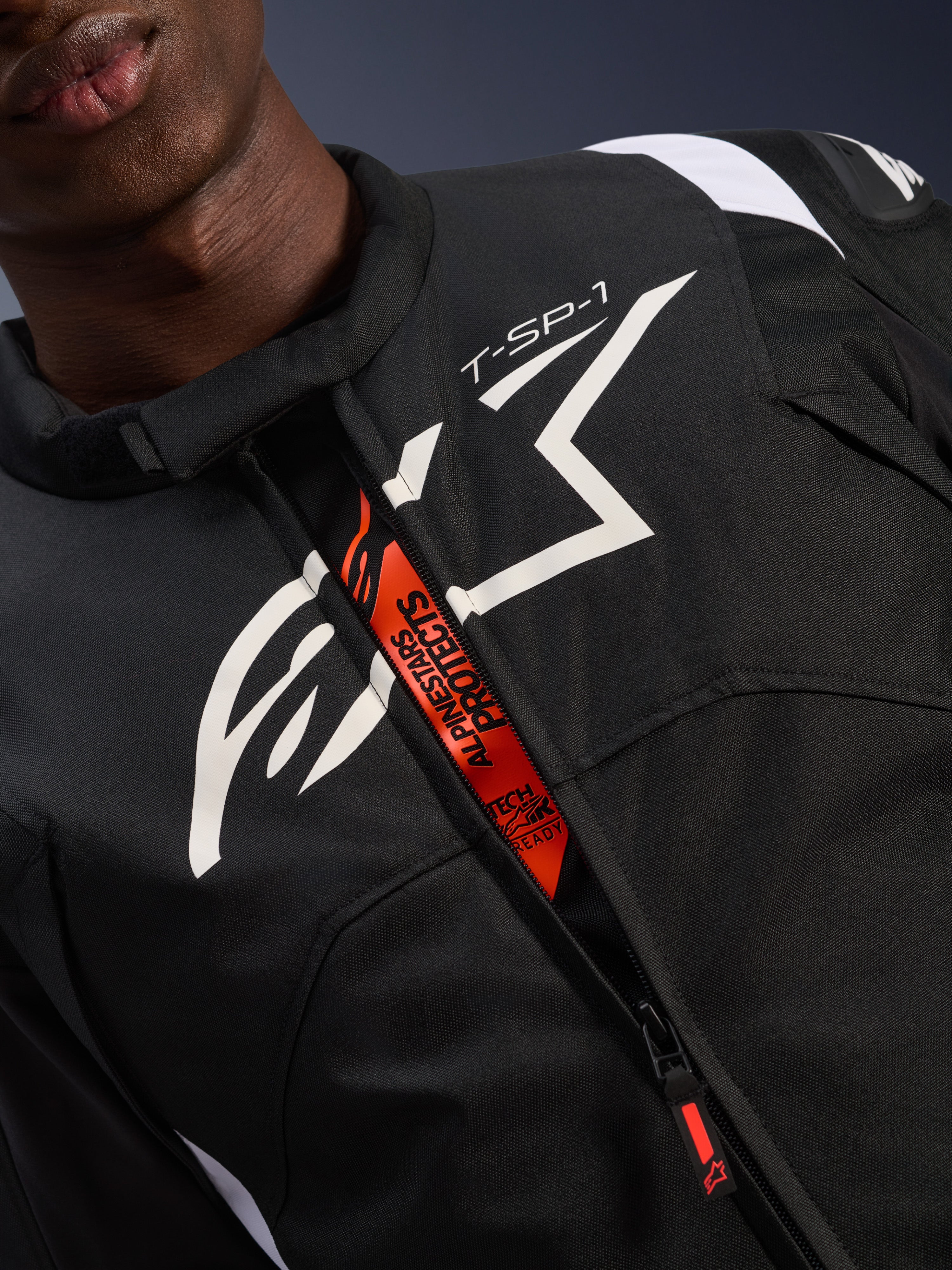 Giacca impermeabile Alpinestars T-SP 1 V2, nero e bianco, con un grande logo bianco sul petto e la scritta 'T-SP-1', patta interna arancione con branding 'Alpinestars Protects' e 'Tech-Air Ready', abbigliamento tecnico da moto in tessuto resistente