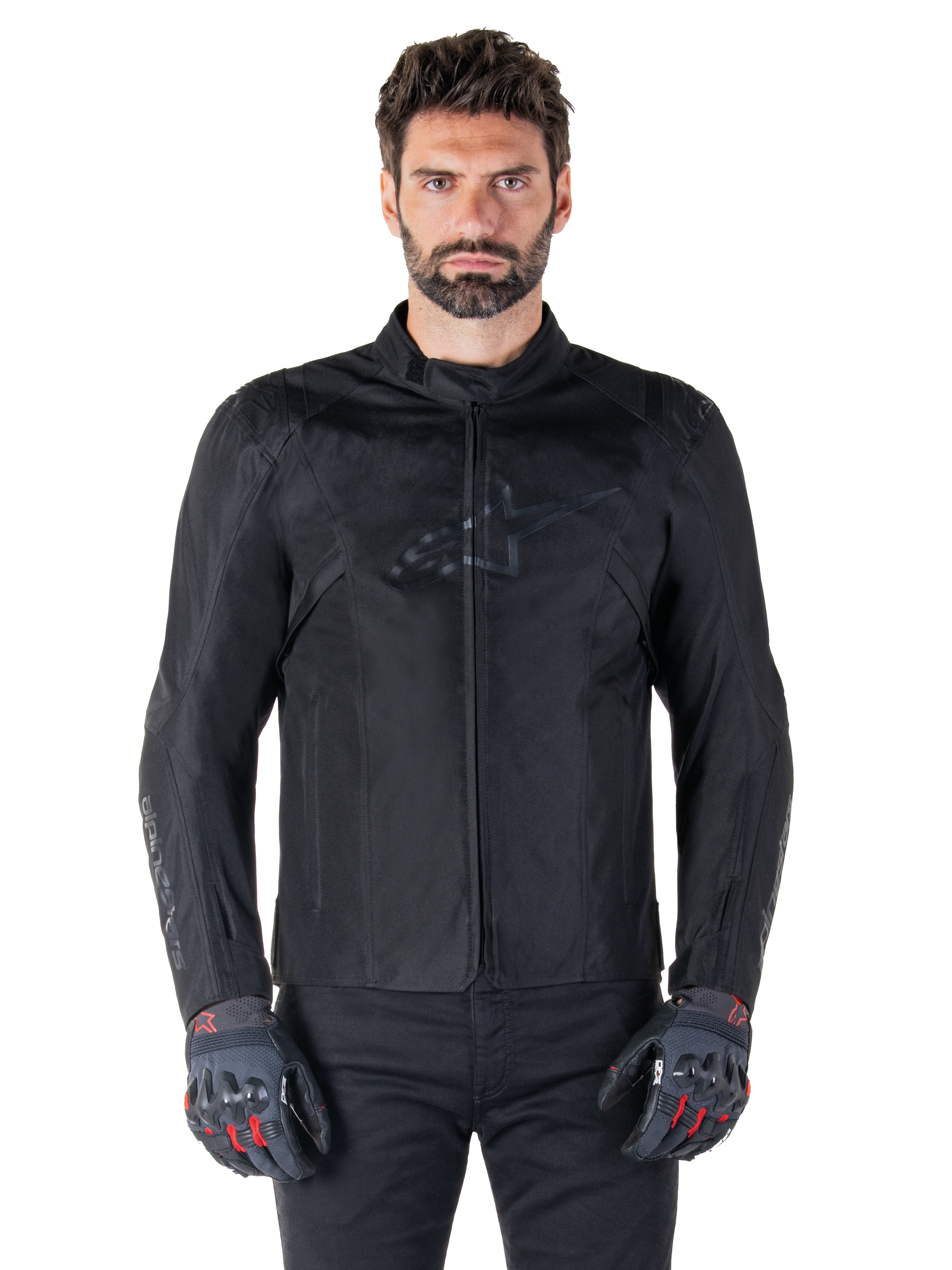 Giacca Alpinestars T-SPS V2 Waterproof, giacca da moto in tessuto nero, indossata da un pilota con guanti neri e rossi, dotata di colletto alto, zip frontale e logo discreto sul petto, protezione impermeabile per guida sportiva e urbana