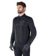 Giacca Alpinestars T-SPS V2 Waterproof, nera, indossata con pantaloni neri e guanti da moto grigi con accenti rossi, con logo nero discreto sul petto e branding sulla manica, giacca in tessuto dal taglio sportivo per guida su strada