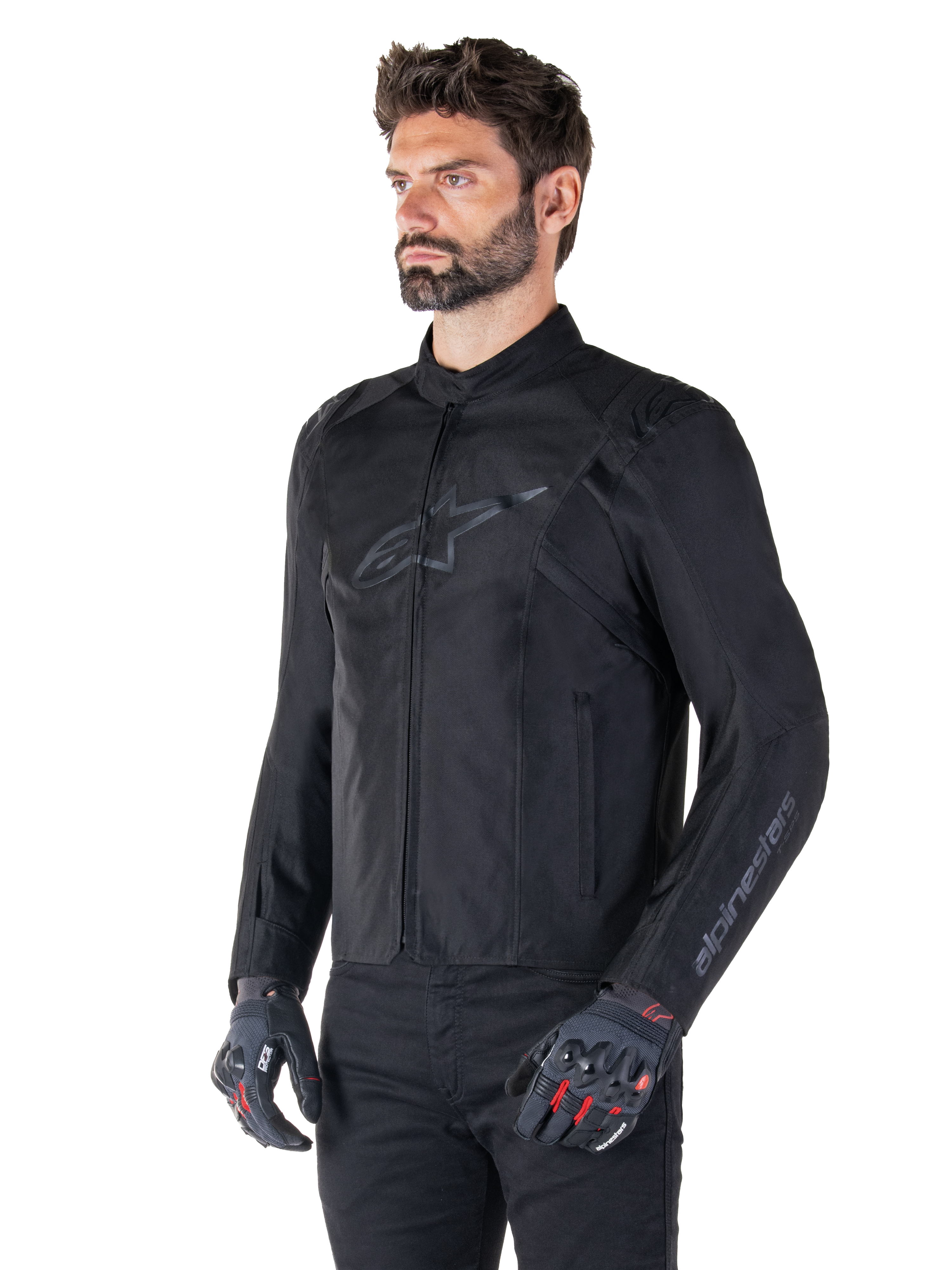 Giacca Alpinestars T-SPS V2 Waterproof, nera, indossata con pantaloni neri e guanti da moto grigi con accenti rossi, con logo nero discreto sul petto e branding sulla manica, giacca in tessuto dal taglio sportivo per guida su strada
