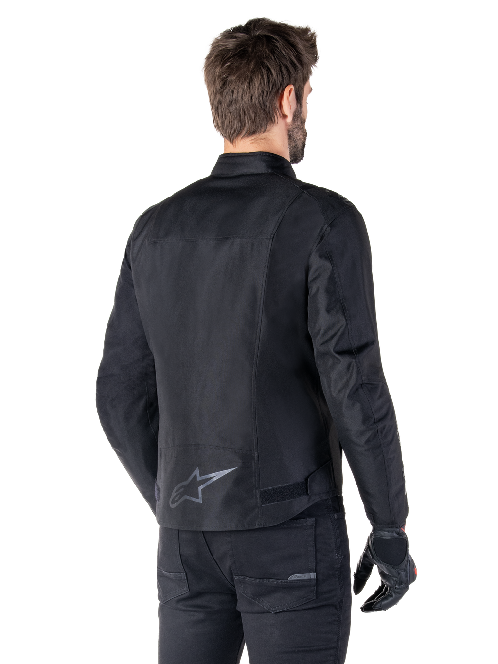 Giacca impermeabile Alpinestars T-SPS V2, nero, vista posteriore su un modello, con logo a stella riflettente sulla parte bassa della schiena, profilo posteriore allungato e cinghie di regolazione in vita, abbinata a pantaloni e guanti da moto neri