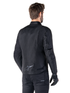 Giacca impermeabile Alpinestars T-SPS V2, nero, vista posteriore su un modello, con logo a stella riflettente sulla parte bassa della schiena, profilo posteriore allungato e cinghie di regolazione in vita, abbinata a pantaloni e guanti da moto neri
