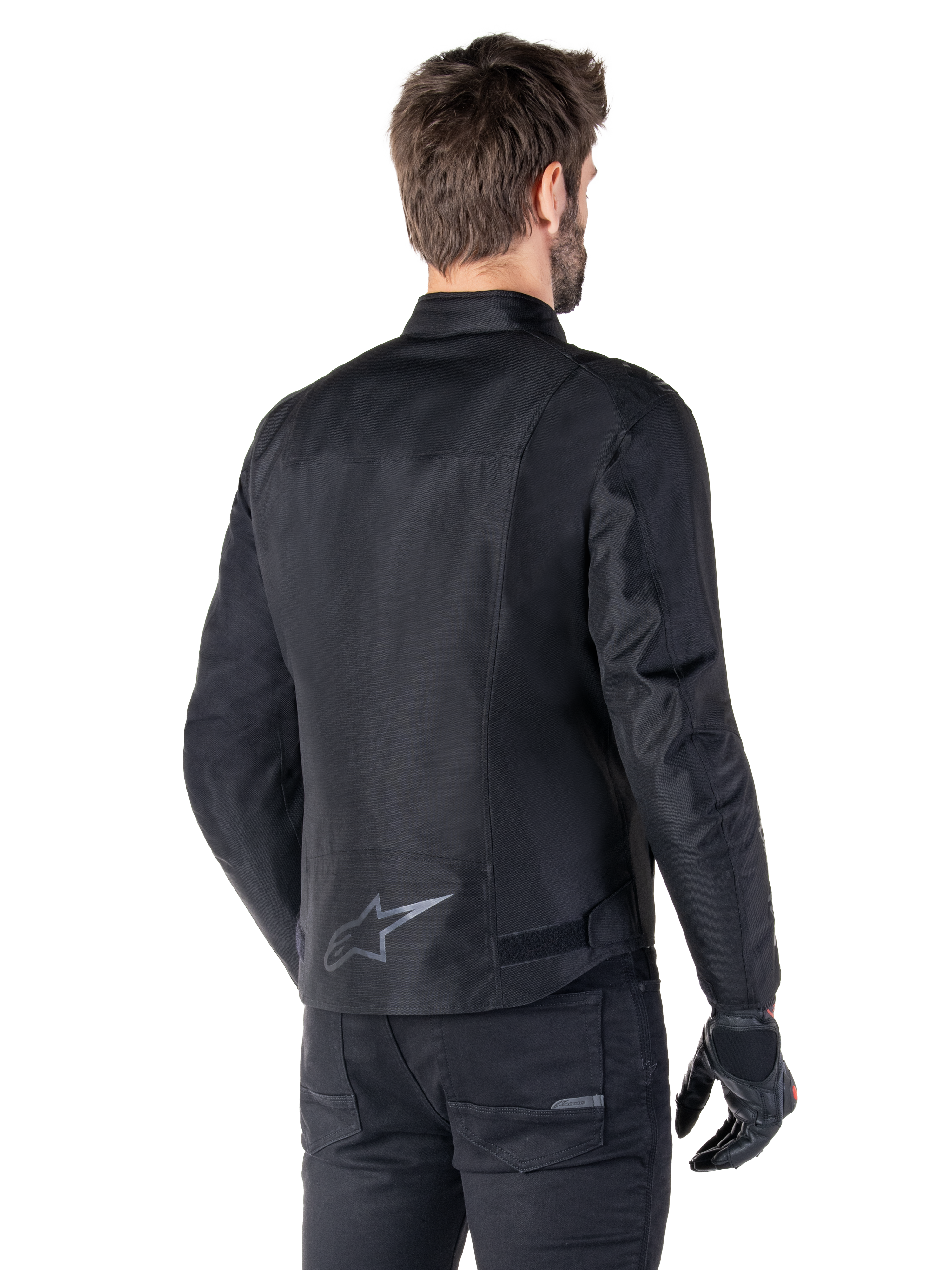 Giacca impermeabile Alpinestars T-SPS V2, nero, vista posteriore su un modello, con logo a stella riflettente sulla parte bassa della schiena, profilo posteriore allungato e cinghie di regolazione in vita, abbinata a pantaloni e guanti da moto neri