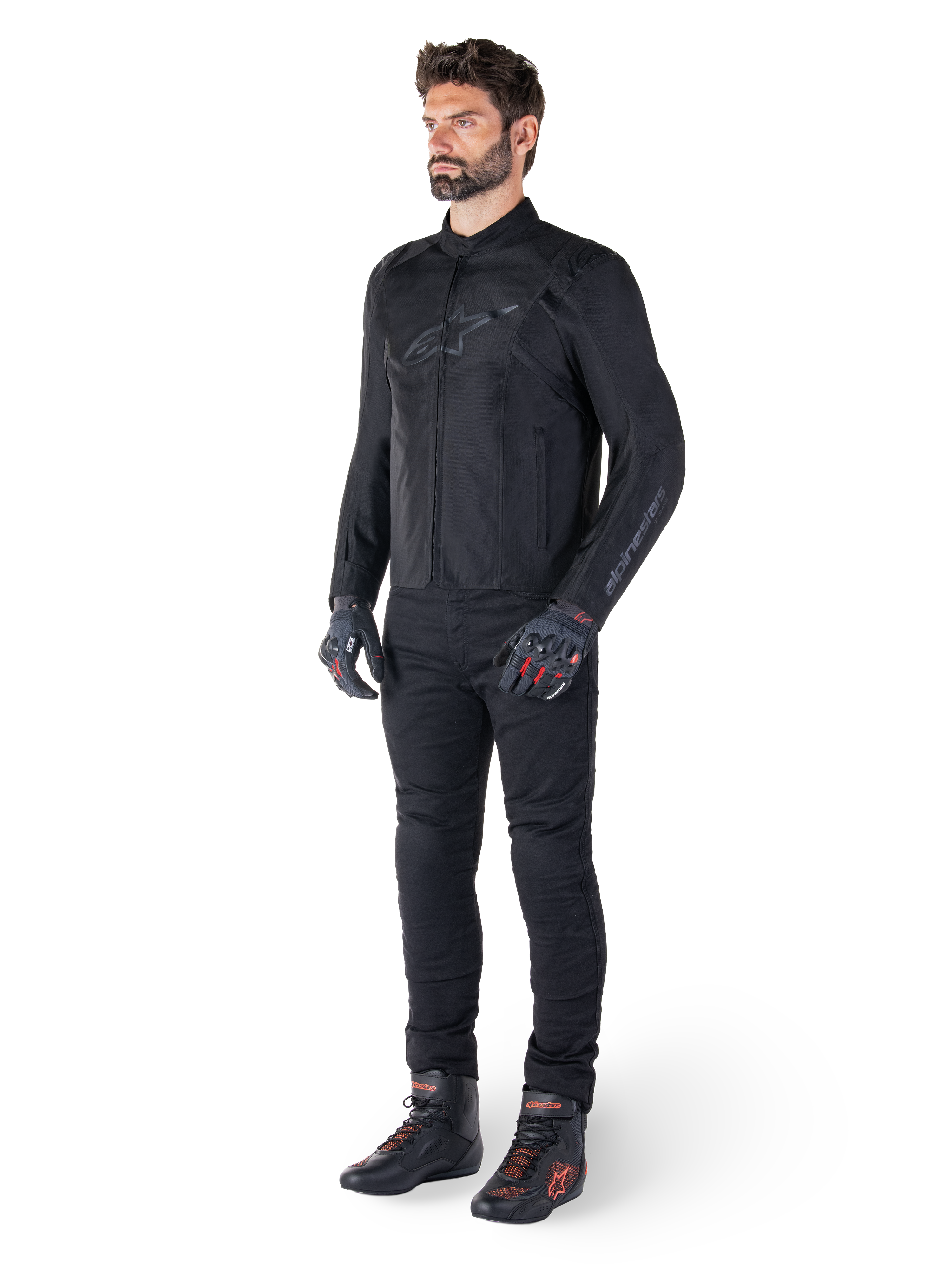 Giacca impermeabile Alpinestars T-SPS V2, nero, indossata da un modello con pantaloni neri coordinati, guanti neri con dettagli rossi e scarpe da moto nere, equipaggiamento completo da moto per guida urbana e sportiva, sfondo bianco