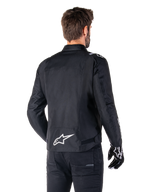 Giacca Alpinestars T-SPS V2 Waterproof, nero e bianco, vista posteriore di un motociclista che indossa la giacca, logo Alpinestars bianco sulla parte bassa della schiena, struttura in tessuto con taglio sportivo, indossata con pantaloni da moto e guanti neri