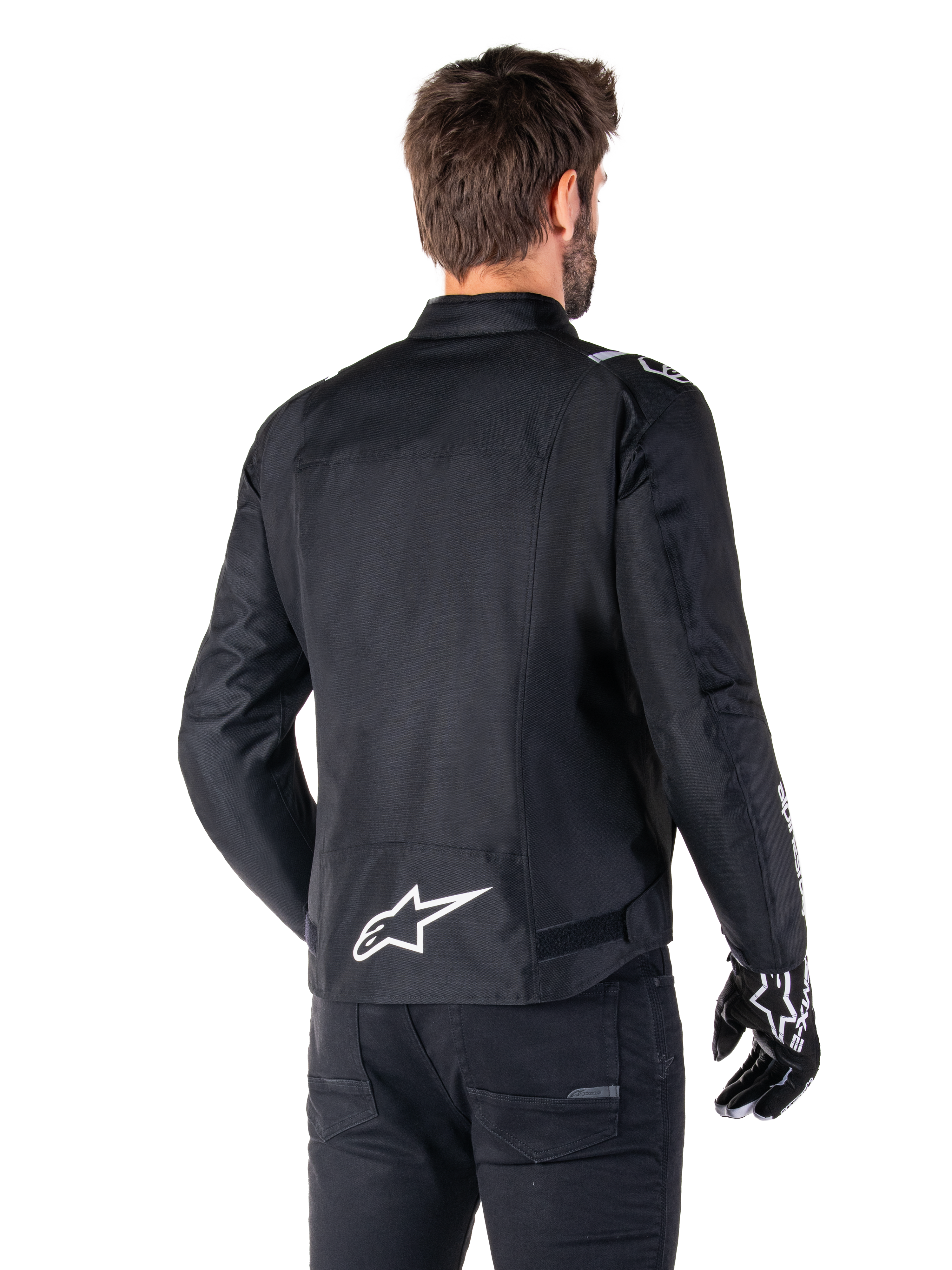 Giacca Alpinestars T-SPS V2 Waterproof, nero e bianco, vista posteriore di un motociclista che indossa la giacca, logo Alpinestars bianco sulla parte bassa della schiena, struttura in tessuto con taglio sportivo, indossata con pantaloni da moto e guanti neri