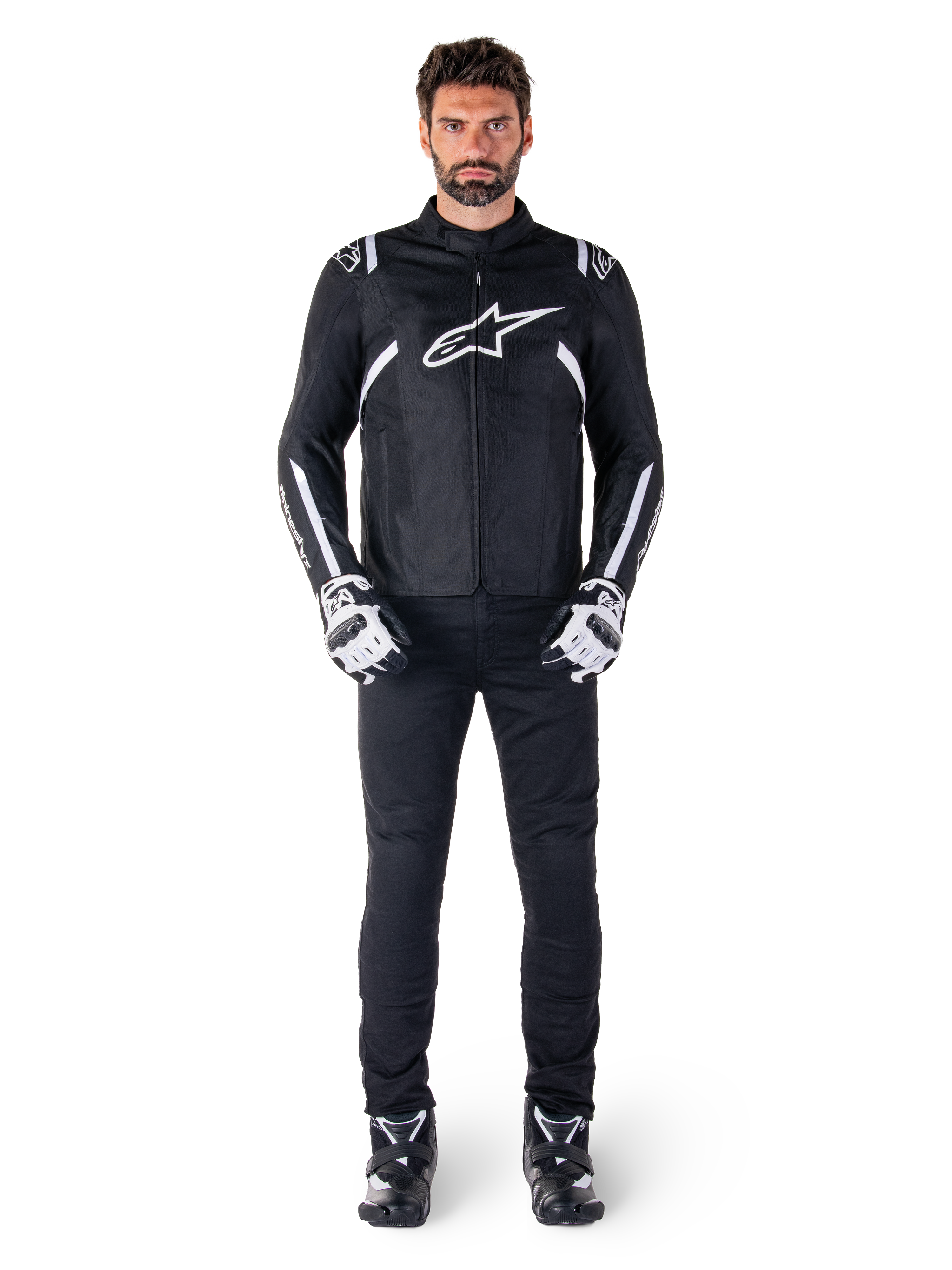 Giacca impermeabile Alpinestars T-SPS V2, nero e bianco, indossata da un modello uomo con pantaloni neri e guanti coordinati, con logo Alpinestars bianco sul petto e dettagli sulle maniche, silhouette dal taglio sportivo per la guida in moto in ogni condizione atmosferica