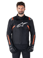 Giacca impermeabile Alpinestars T-SPS V2, nero, bianco e rosso fluo, indossata da un modello con guanti da moto neri, caratterizzata da un grande logo Alpinestars bianco sul petto e vivaci dettagli arancioni sulle spalle e sulle maniche.