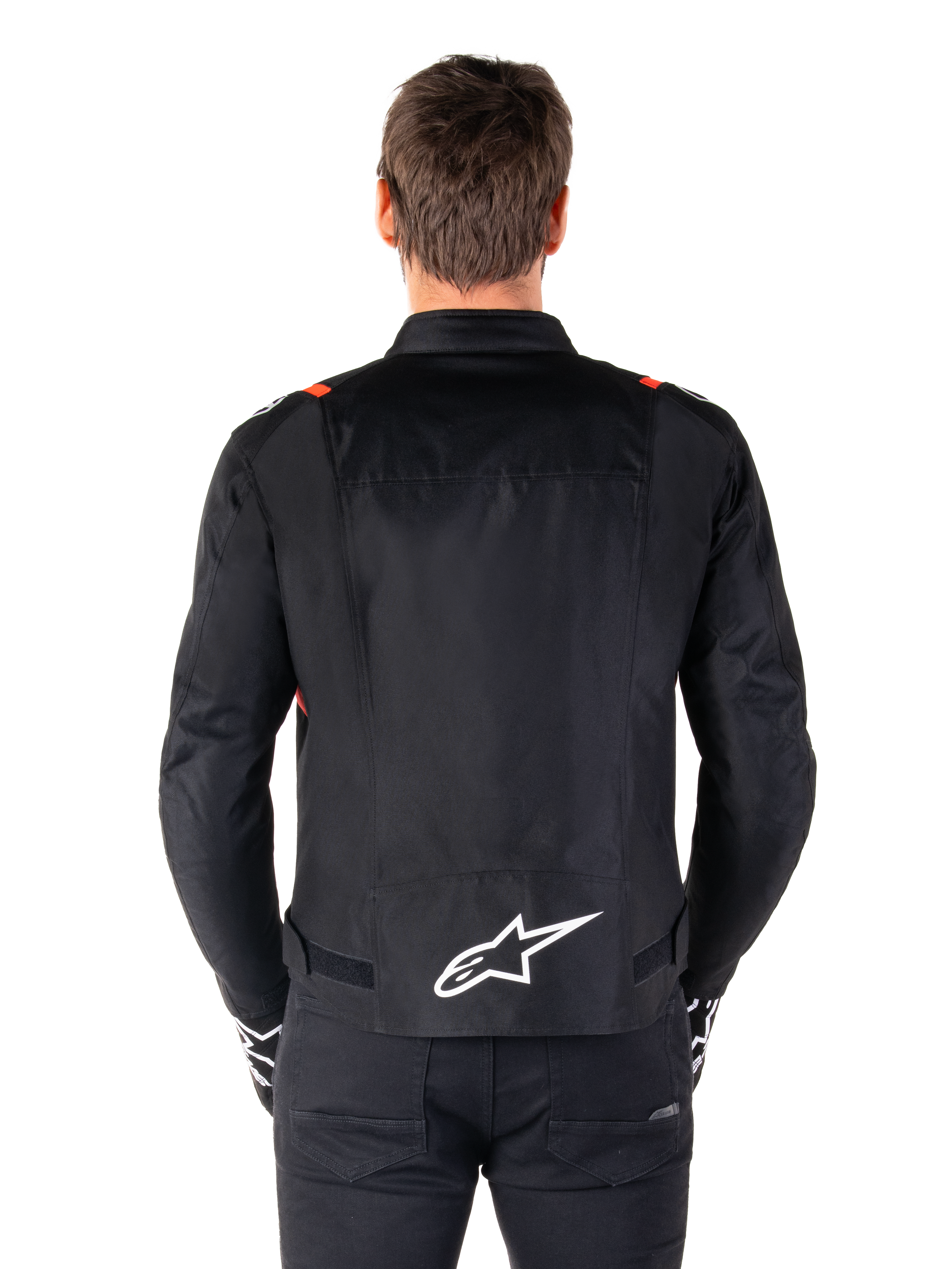 Giacca Alpinestars T-SPS V2 Waterproof, nero con accenti bianco e rosso fluo, vista posteriore con grande logo Alpinestars bianco sulla parte bassa della schiena, giacca da moto in tessuto dal taglio sportivo, indossata con pantaloni e guanti neri