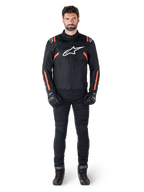 Modello che indossa la giacca Alpinestars T-SPS V2 Waterproof in Nero Bianco Rosso Fluo, abbinata a pantaloni da moto e guanti neri, con logo Alpinestars bianco sul petto e dettagli rossi sulle maniche.