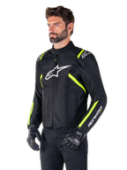 Modello che indossa la giacca impermeabile Alpinestars T-SPS V2, nero con dettagli bianco e giallo fluo, con logo Astars bianco sul petto e strisce giallo fluorescente su spalle e maniche, abbinata a pantaloni neri e guanti da guida, giacca da moto in tessuto dal taglio sportivo.