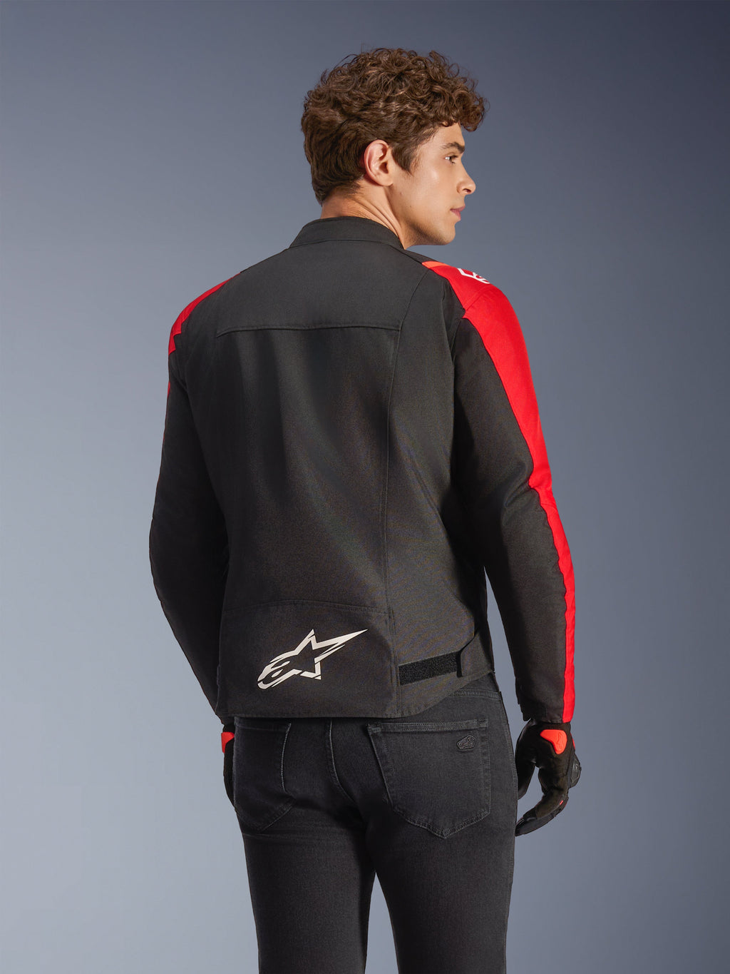 Giacca impermeabile Alpinestars T-SPS V2 in nero e rosso, vista posteriore su un modello, con logo Alpinestars bianco sulla parte bassa della schiena, pannelli delle maniche in rosso vivace e silhouette dal taglio sportivo, indossata con guanti neri e jeans.