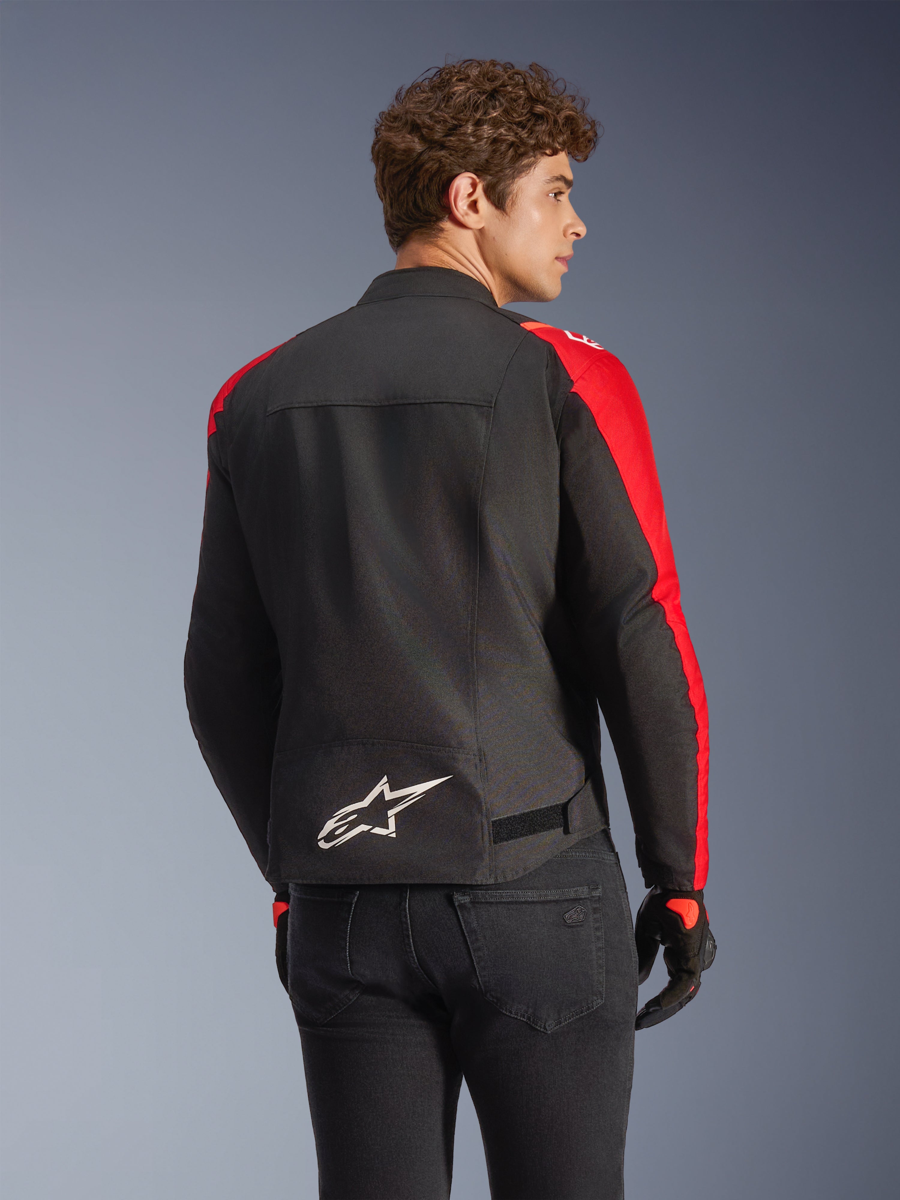 Giacca impermeabile Alpinestars T-SPS V2 in nero e rosso, vista posteriore su un modello, con logo Alpinestars bianco sulla parte bassa della schiena, pannelli delle maniche in rosso vivace e silhouette dal taglio sportivo, indossata con guanti neri e jeans.