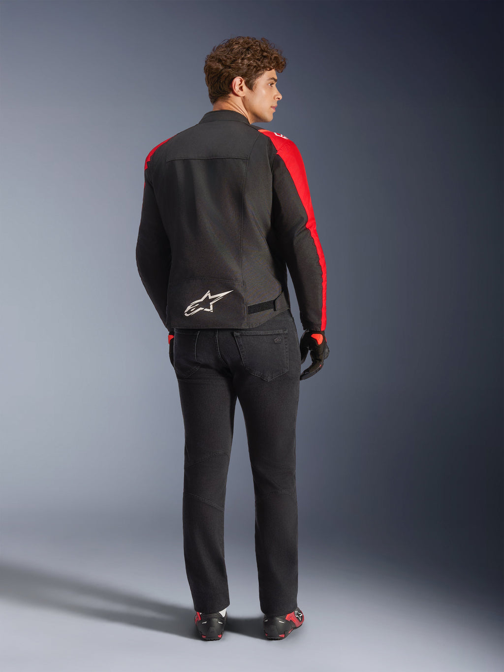 Giacca impermeabile Alpinestars T-SPS V2, nero rosso rosso fluo, vista posteriore su modello, logo Alpinestars bianco sulla parte bassa della schiena, indossata con pantaloni da moto neri e guanti, giacca sportiva da moto in tessuto impermeabile