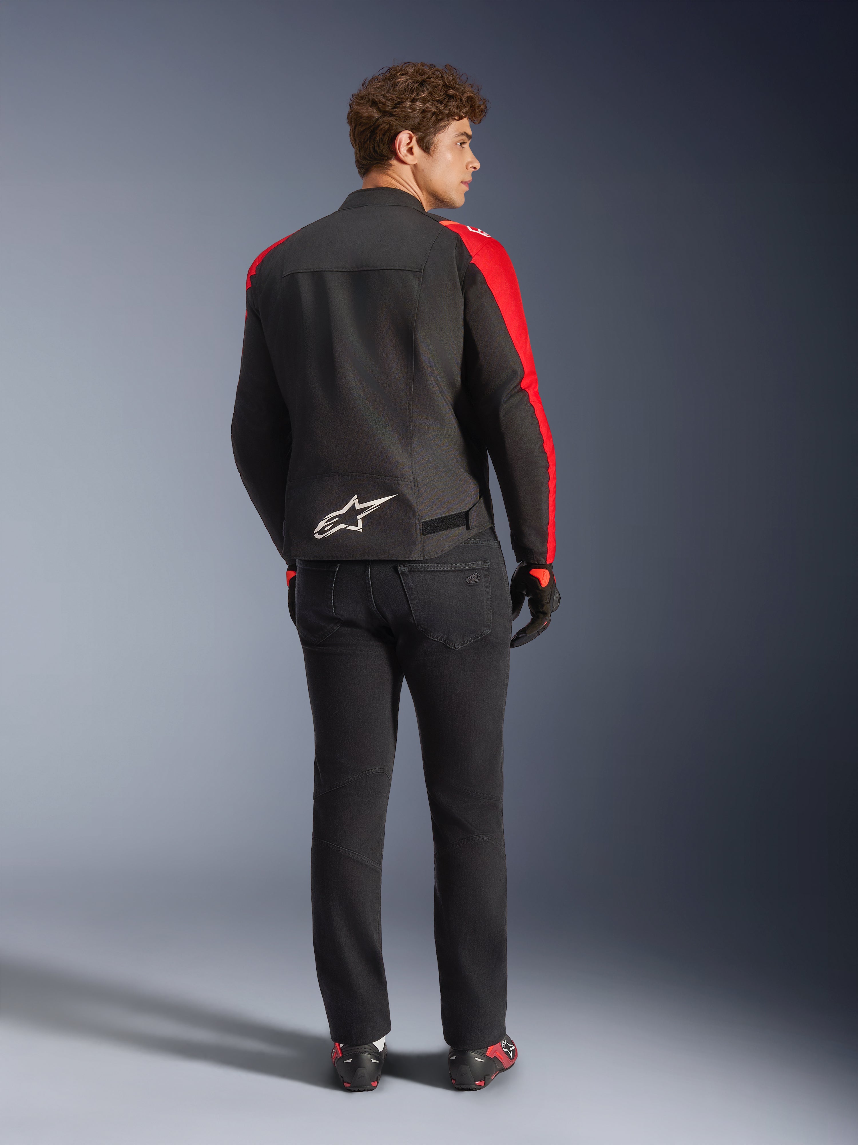 Giacca impermeabile Alpinestars T-SPS V2, nero rosso rosso fluo, vista posteriore su modello, logo Alpinestars bianco sulla parte bassa della schiena, indossata con pantaloni da moto neri e guanti, giacca sportiva da moto in tessuto impermeabile