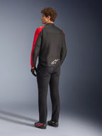 Giacca impermeabile Alpinestars T-SPS V2 in nero e rosso fluo, vista posteriore su modello, indossata con pantaloni da moto neri e scarpe tecniche, con logo Alpinestars bianco sulla parte bassa della schiena, design in tessuto dal taglio sportivo
