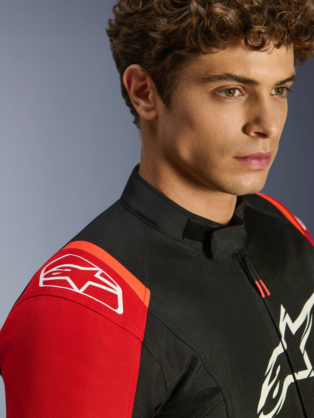 Giacca impermeabile Alpinestars T-SPS V2, giacca da moto in tessuto, nero, rosso e rosso fluo, primo piano del modello che indossa la giacca, con prominenti loghi Alpinestars bianchi su spalla e petto, colletto a basso profilo e vestibilità sportiva.
