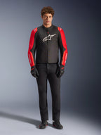 Modello che indossa la giacca Alpinestars T-SPS V2 Waterproof, nero con maniche rosse e rosso fluo, logo bianco sul petto, abbinata a pantaloni neri, guanti e scarpe da moto neri, design sportivo per abbigliamento moto stradale