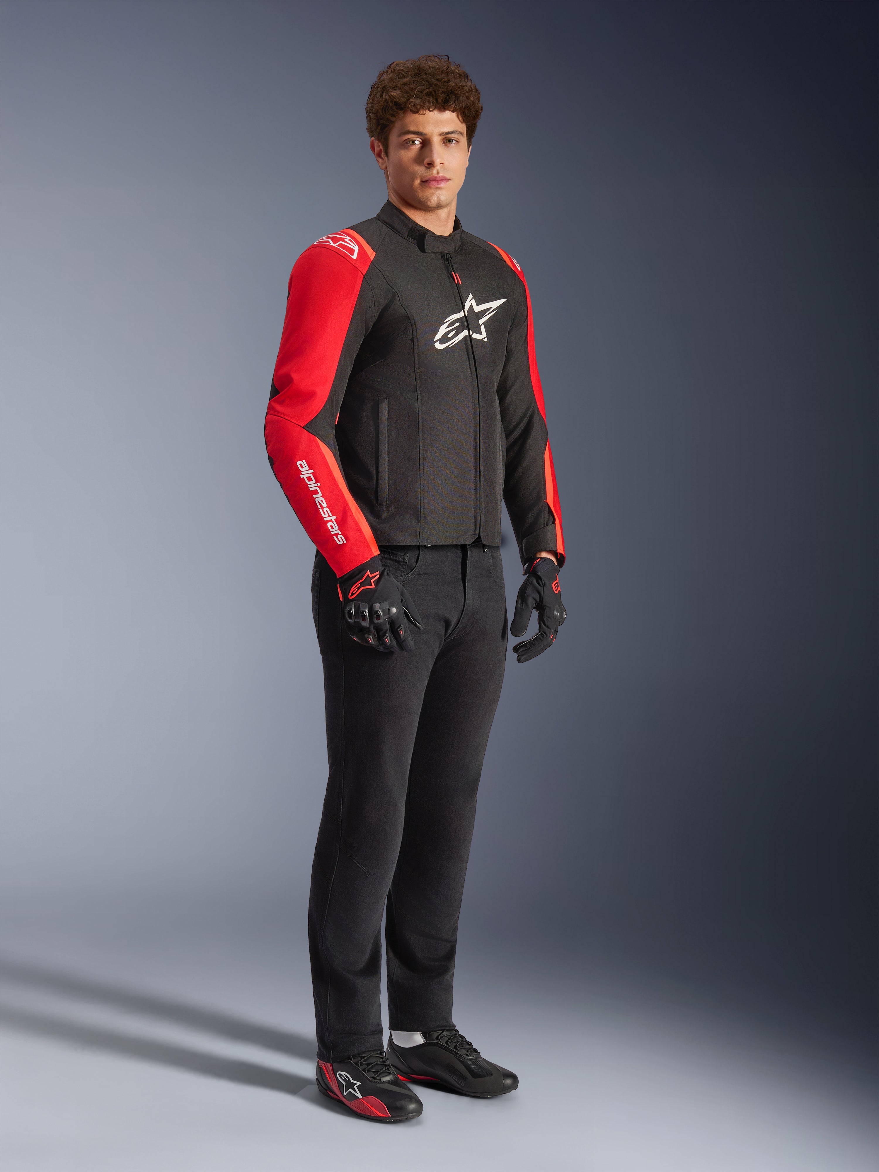 Modello maschile con giacca impermeabile Alpinestars T-SPS V2 in nero e rosso fluo, maniche rosse con logo bianco e grande logo sul petto, abbinata a pantaloni da moto neri, guanti e scarpe tecniche a profilo basso, vista in studio a figura intera.