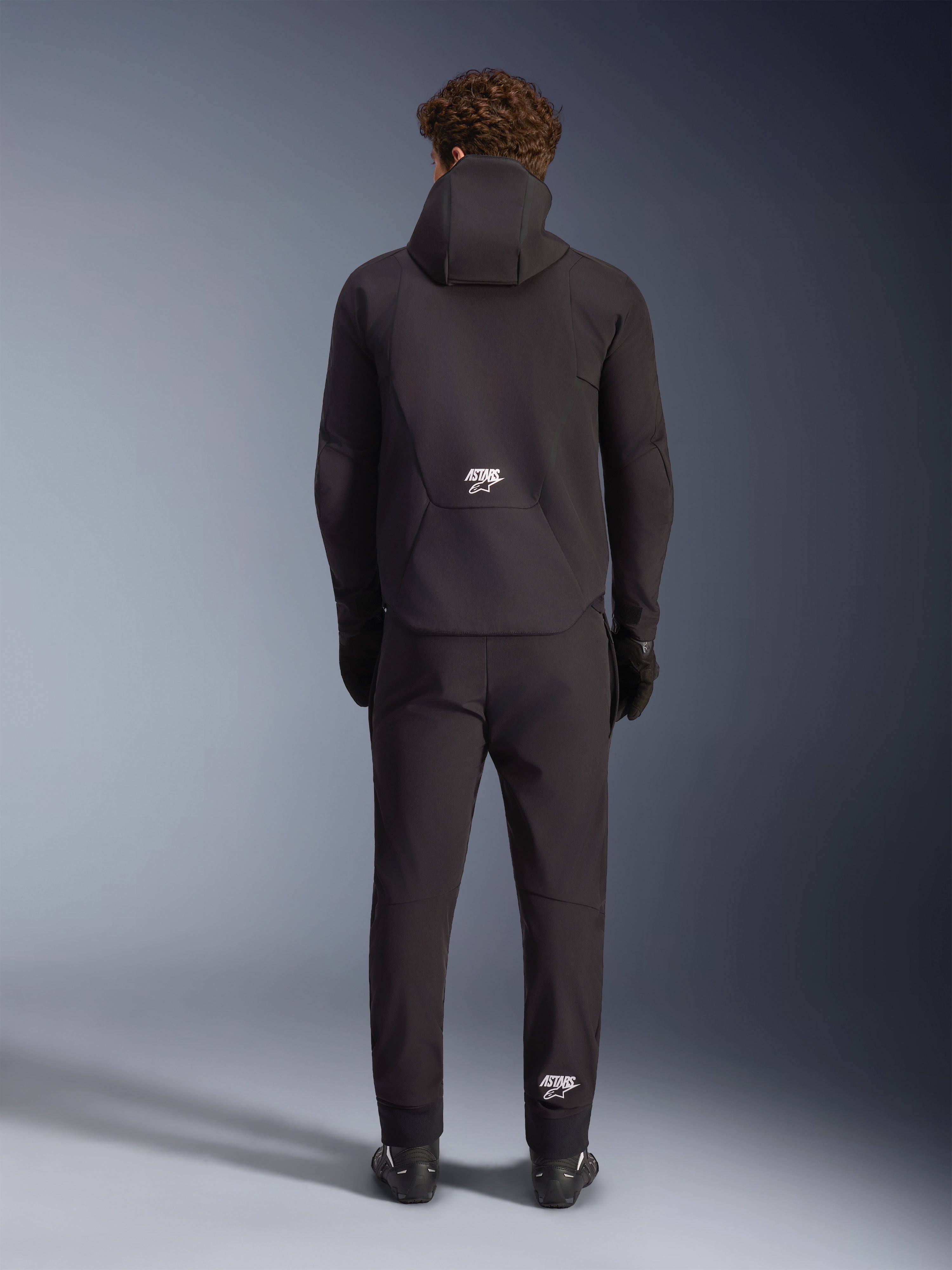 Giacca Alpinestars Aeroshell Waterproof, nera, vista dal retro su un modello con pantaloni e guanti da moto neri coordinati, design softshell sportivo con cappuccio e loghi ASTARS bianchi, abbigliamento da moto moderno ed elegante