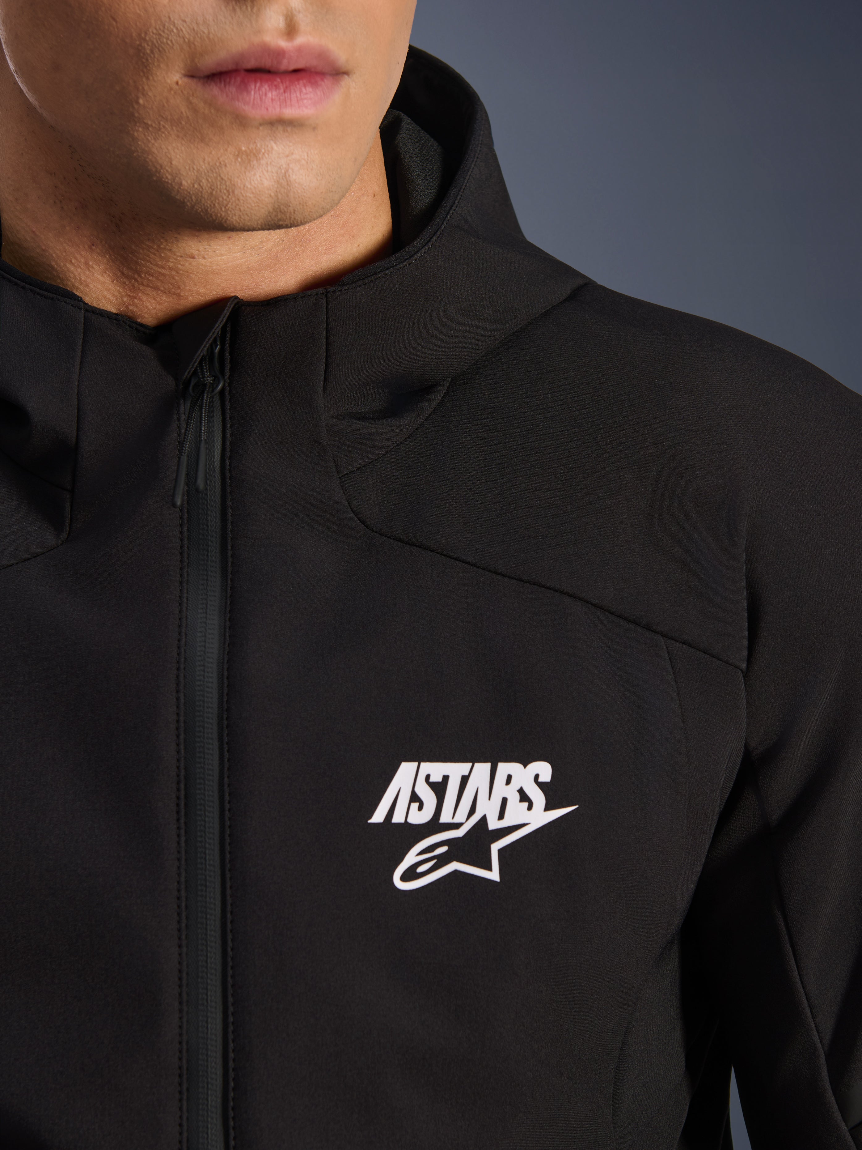 Alpinestars Aeroshell Waterproof Jacket, giacca softshell da moto nera, vista ravvicinata del petto con logo a stella ASTARS bianco e cerniera anteriore impermeabile, cappuccio integrato, design sportivo moderno