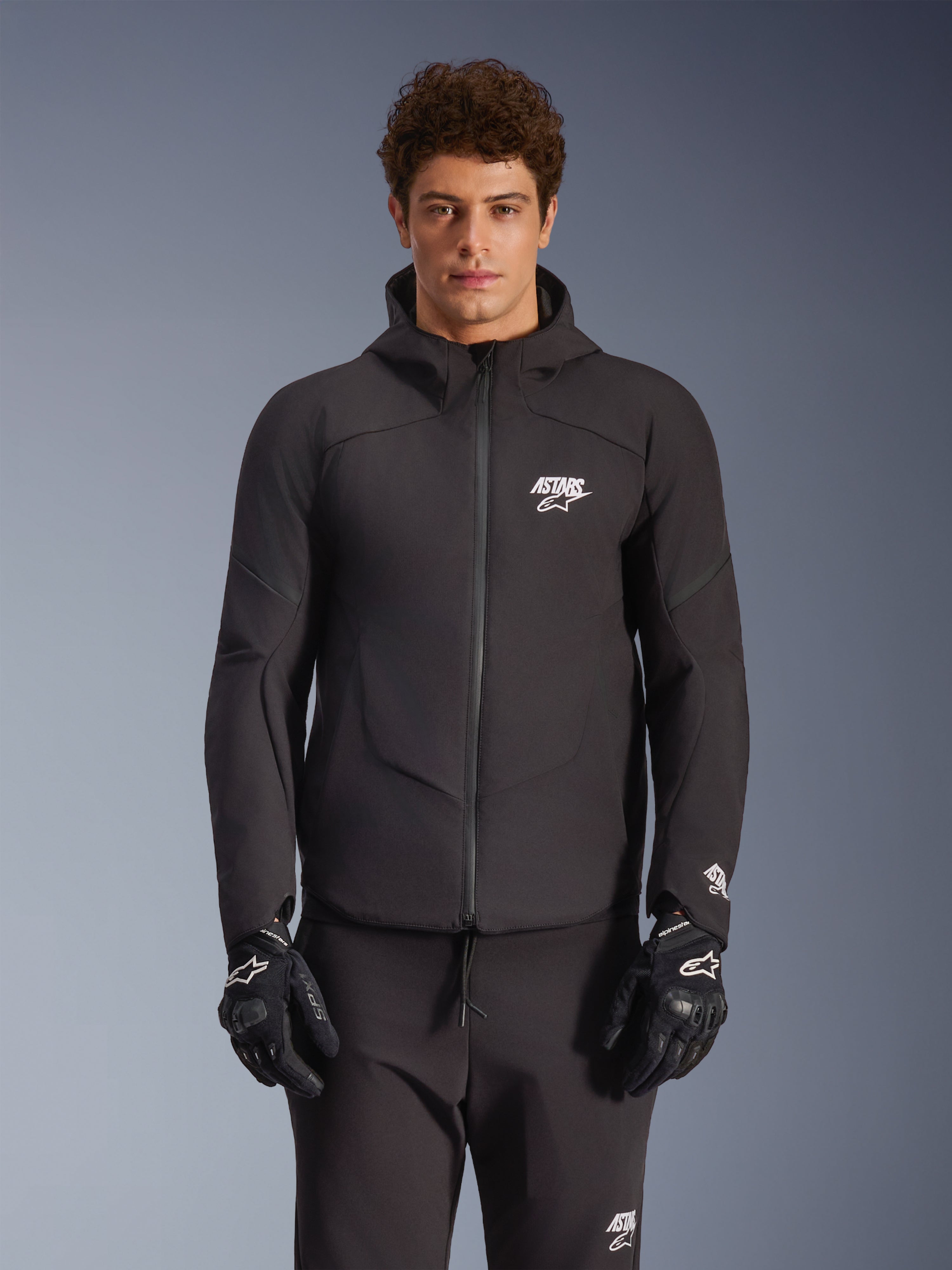Alpinestars Aeroshell Waterproof Jacket, nera, giacca da moto urbana, design con cappuccio e cerniera frontale completa, loghi bianchi su petto e maniche, indossata con pantaloni e guanti neri coordinati