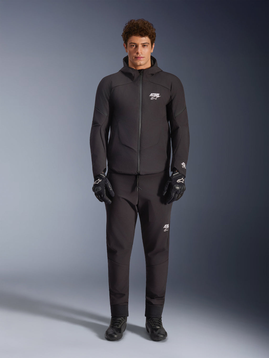 Modello che indossa la giacca Alpinestars Aeroshell Waterproof Jacket in nero, abbinata a pantaloni tecnici neri e guanti da moto coordinati, caratterizzata da un design softshell con cappuccio e accenti con logo bianco, outfit moderno ispirato al motorsport per la guida su strada