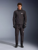 Modello che indossa la giacca Alpinestars Aeroshell Waterproof Jacket in nero, abbinata a pantaloni tecnici neri e guanti da moto coordinati, caratterizzata da un design softshell con cappuccio e accenti con logo bianco, outfit moderno ispirato al motorsport per la guida su strada