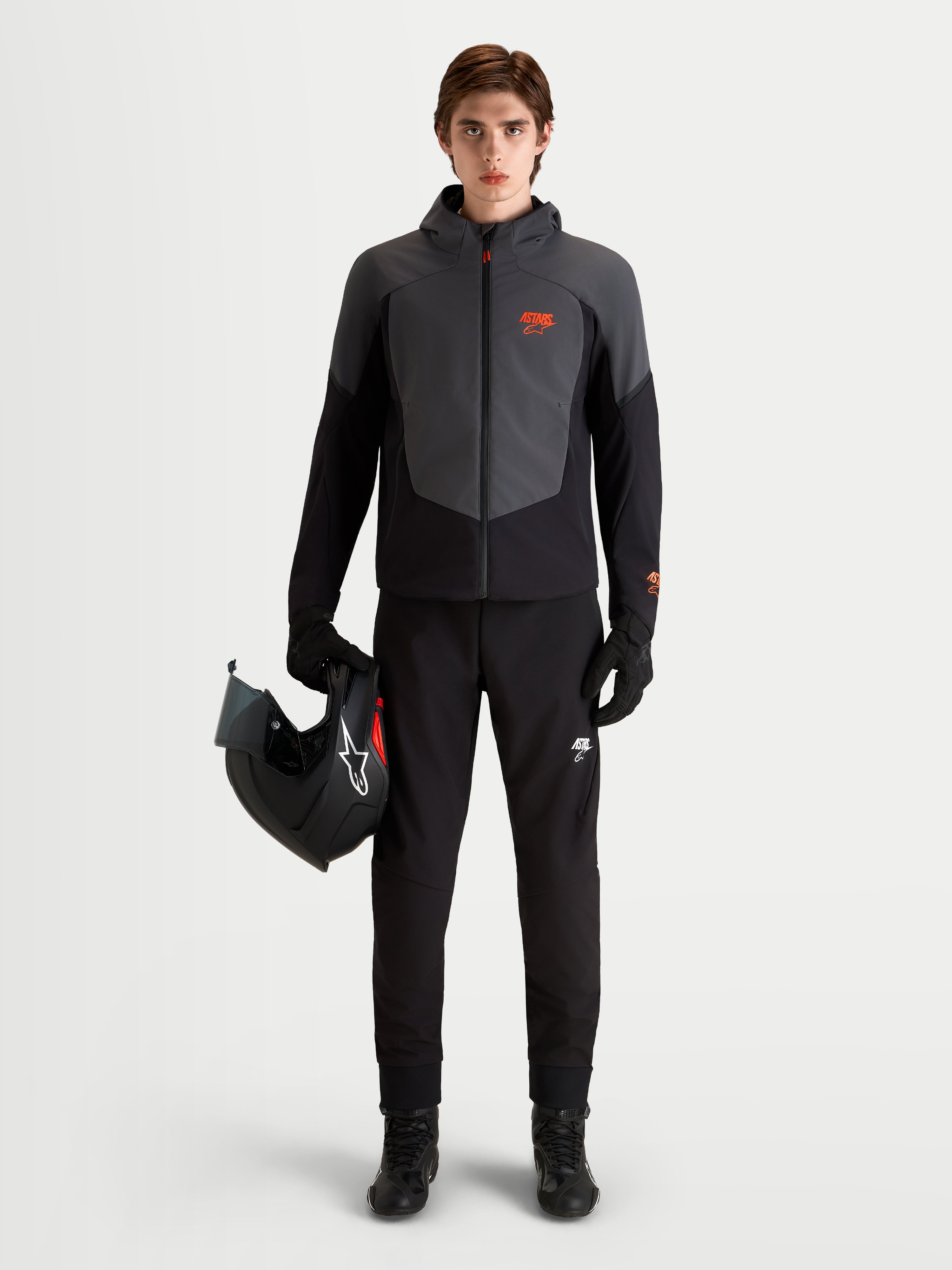 Alpinestars Aeroshell Waterproof Jacket, giacca da moto impermeabile da uomo in nero e grigio scuro con cappuccio integrato, indossata da un modello che tiene un casco da moto
