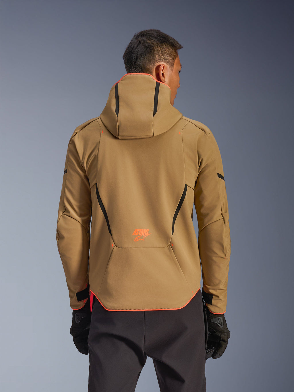 Giacca impermeabile Alpinestars Aeroshell, Dark Khaki e Arancione Fluo, vista posteriore con design a felpa con cappuccio e profili arancioni su cappuccio e orlo, logo arancione sulla parte bassa della schiena, indossata con guanti neri, abbigliamento da moto softshell sportivo crossover.