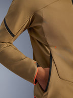 Primo piano della giacca Alpinestars Aeroshell Waterproof, Dark Khaki con accenti neri e arancione fluo, dettaglio della tasca nascosta con zip e del polsino, tessuto softshell elasticizzato per la guida in moto