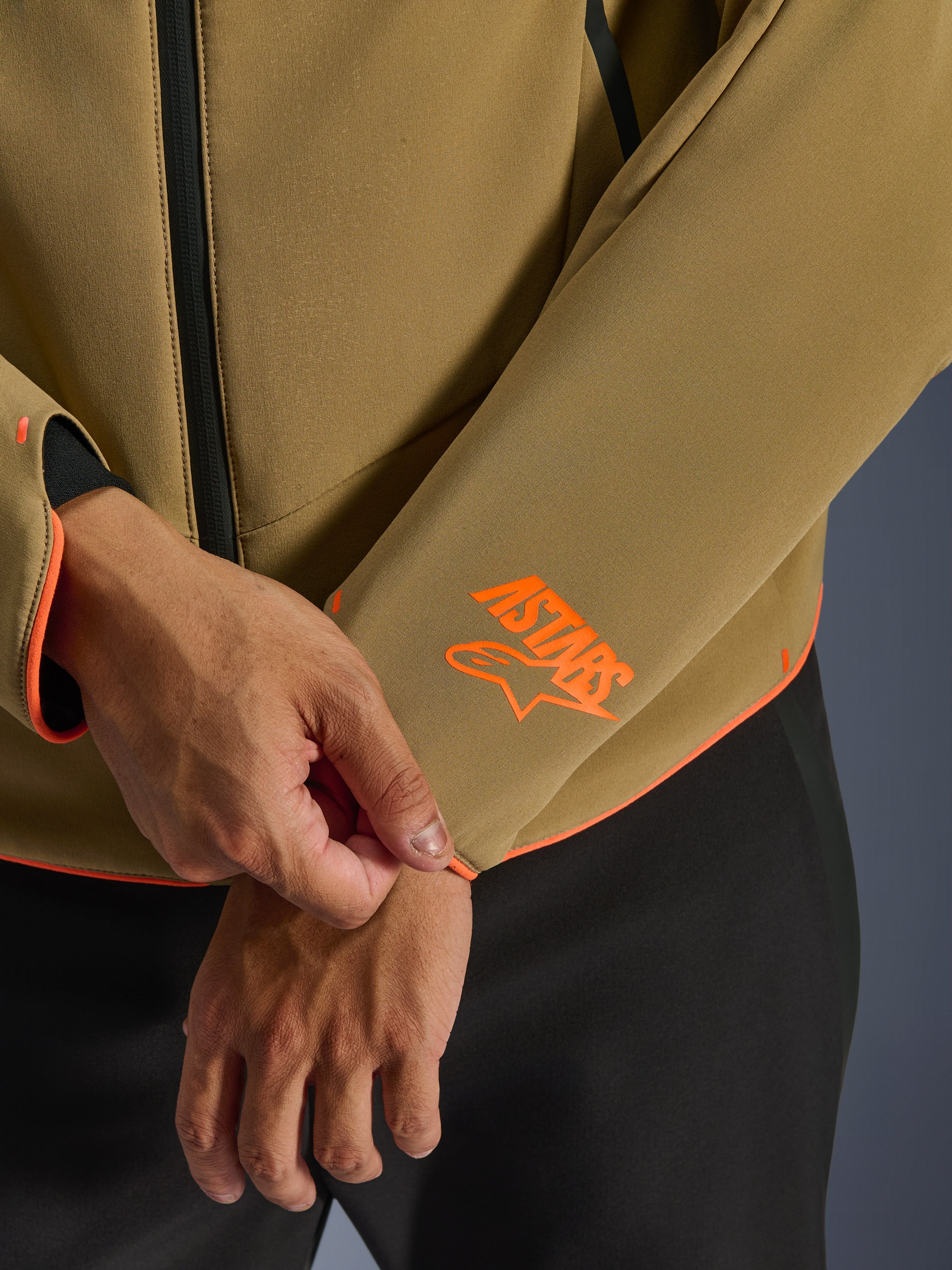 Giacca Alpinestars Aeroshell Waterproof, Dark Khaki con accenti Orange Fluo, primo piano del polsino e dell'orlo inferiore con logo Astars arancione brillante, profili arancione neon e dettaglio della cerniera frontale impermeabile, costruzione softshell sportiva.